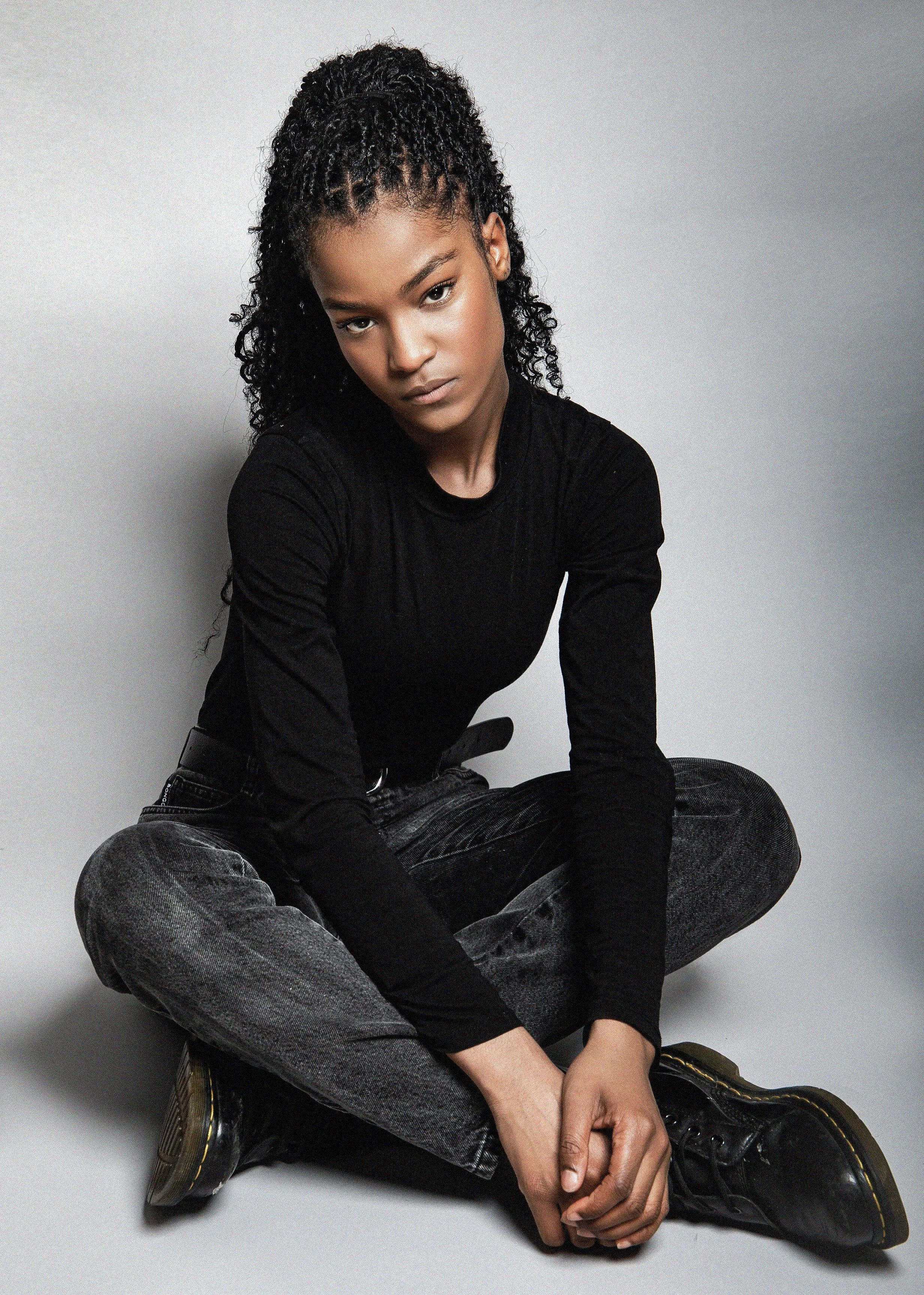 A young woman with curly black hair, wearing a black long-sleeve shirt and gray jeans, sitting cross-legged on the floor against a plain gray background.