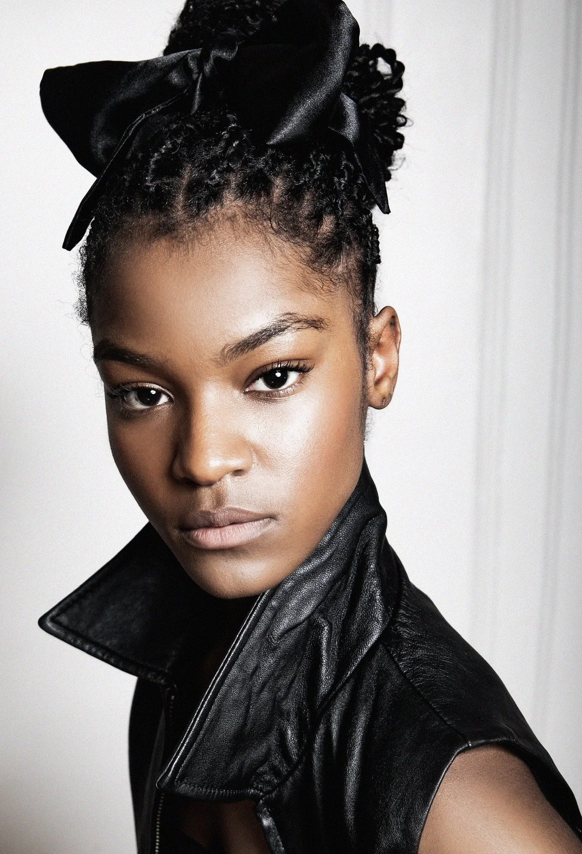 Close-up portrait of a woman with natural hair styled in twists with a large black satin bow on top, wearing a black leather jacket, looking directly at the camera with a neutral expression.