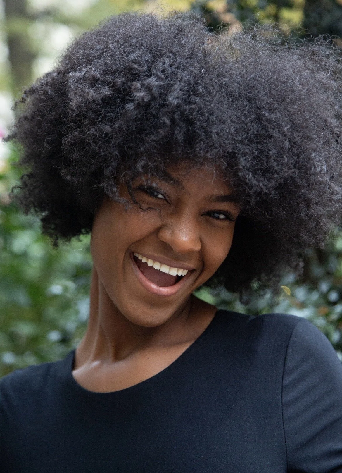 A woman with dark skin and a big afro hairstyle, smiling and laughing outdoors against a green leafy background.