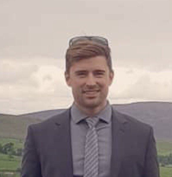 A man in a suit and tie standing outdoors with rolling hills and a cloudy sky in the background.