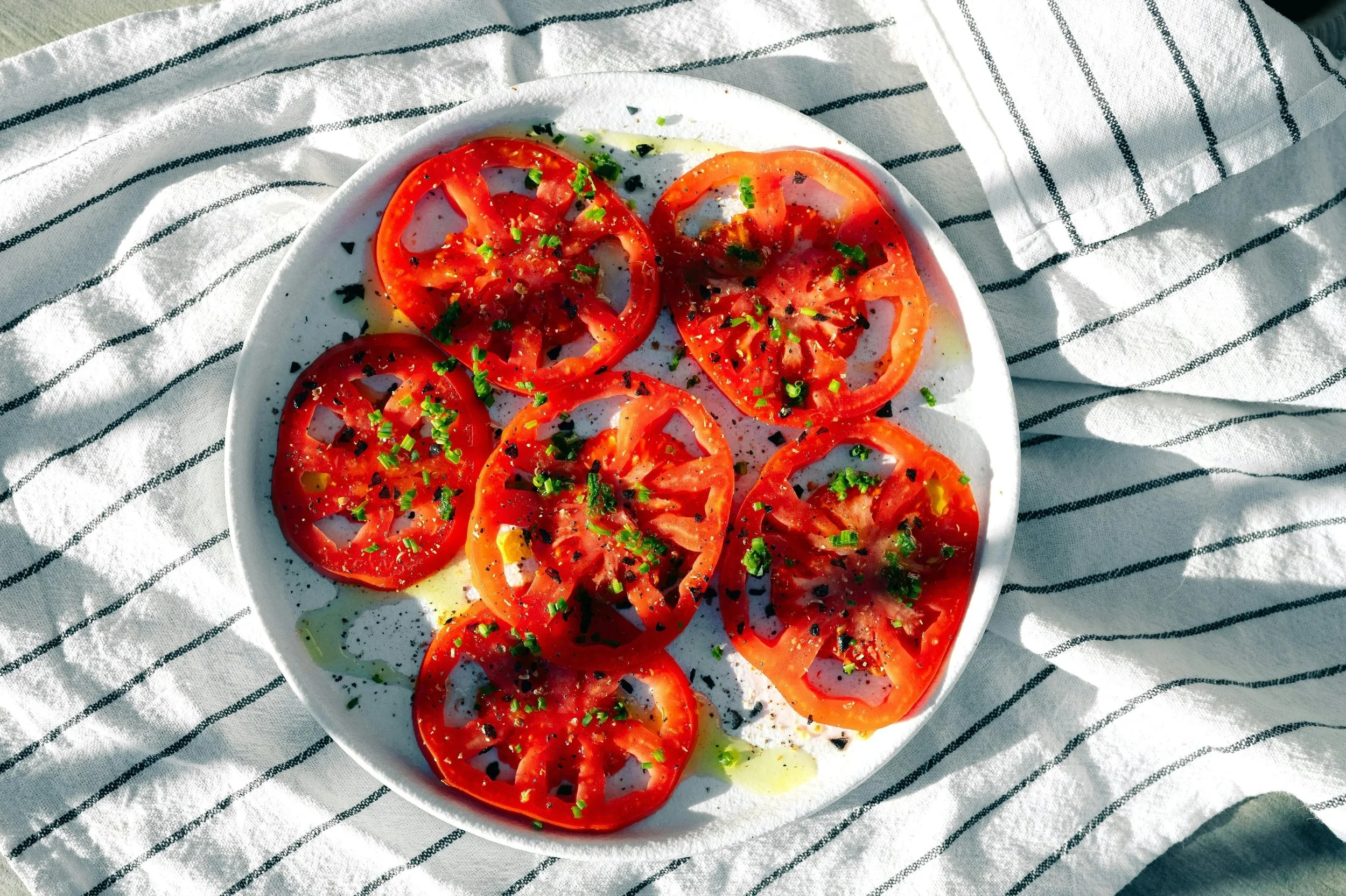 Plate of sliced tomatoes garnished with chopped green herbs and black pepper, placed on a striped cloth.