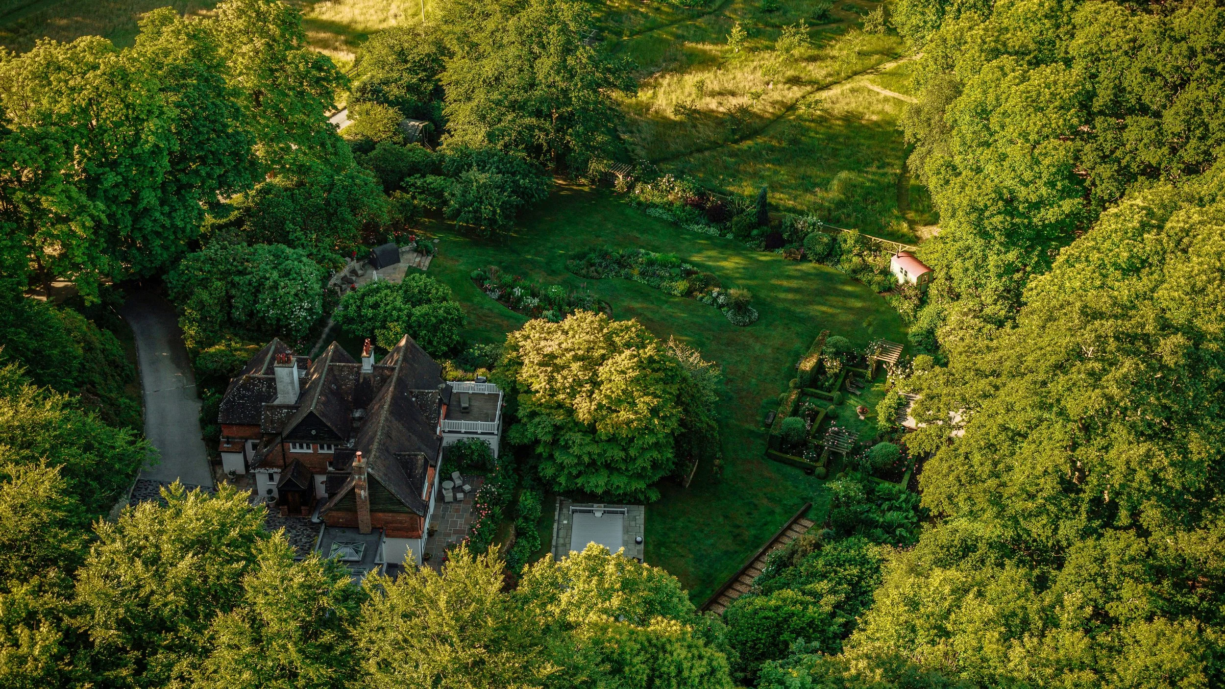 Aerial view of a lush, green backyard garden with trees, flower beds, a patio area, and neighboring houses, surrounded by extensive greenery.