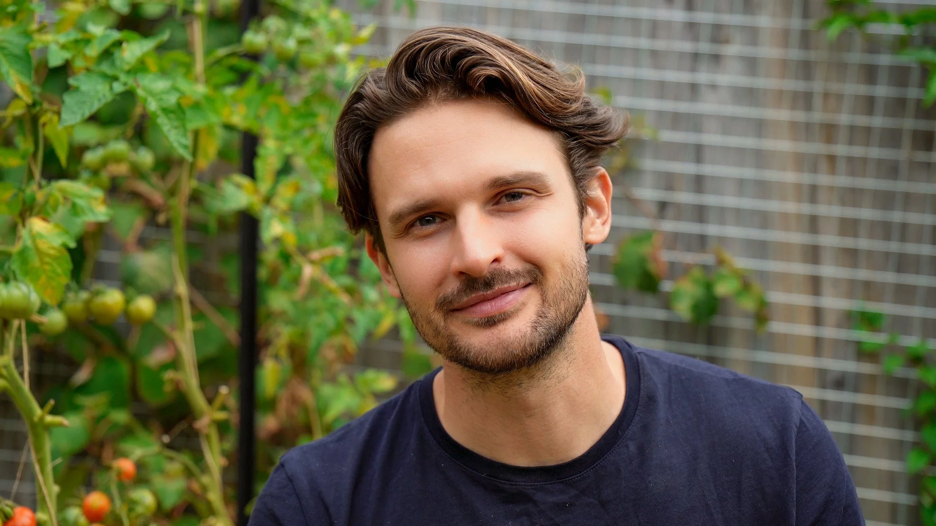 A young man with medium-length brown hair, a beard, and a slight smile stands in a garden with green tomato plants and a metal fence in the background.