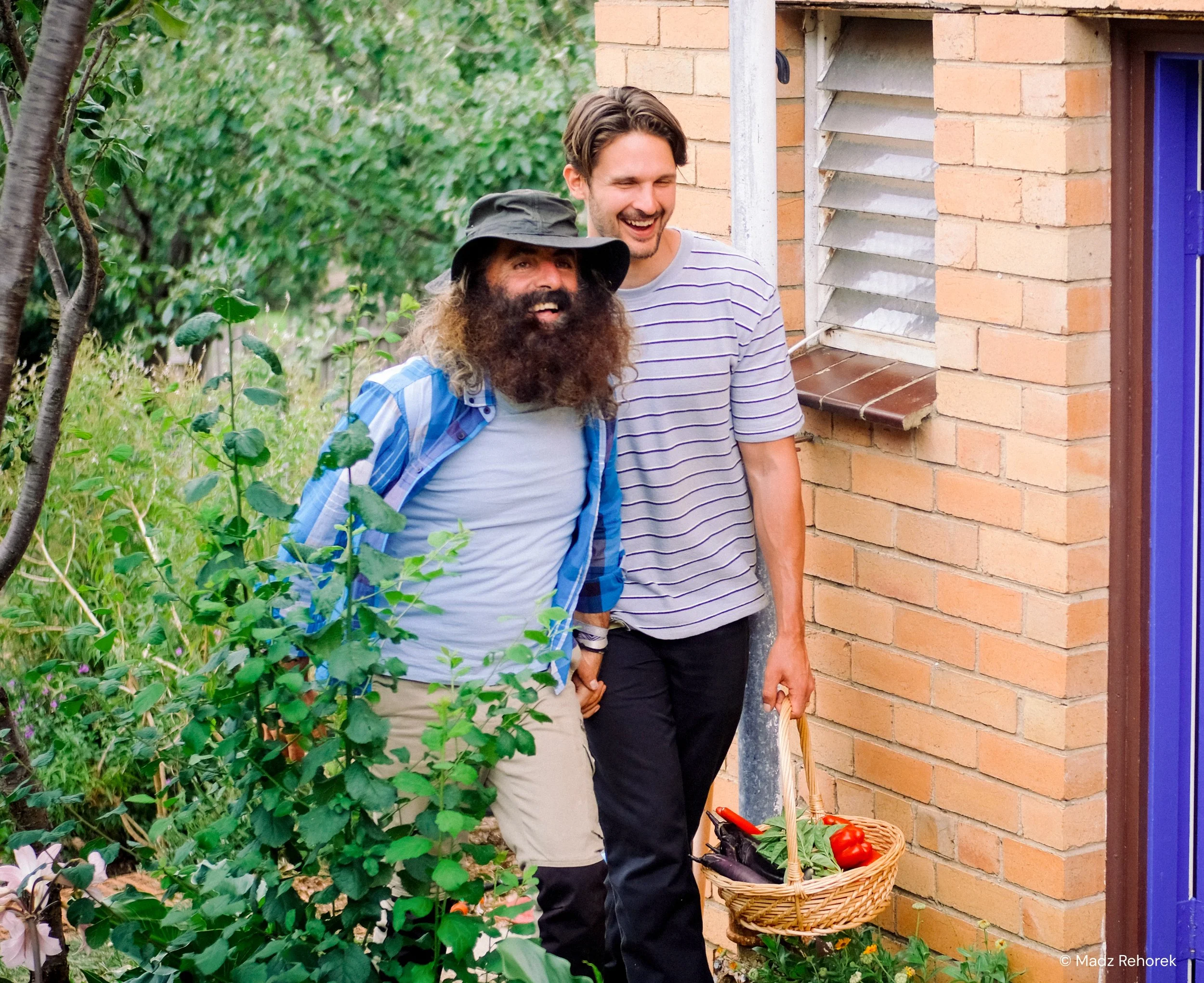 Two men hold hands outside near a brick building, one with a beard, wearing a hat and plaid shirt, and the other clean-shaven, in a striped shirt, standing near a basket of tomatoes and greenery.