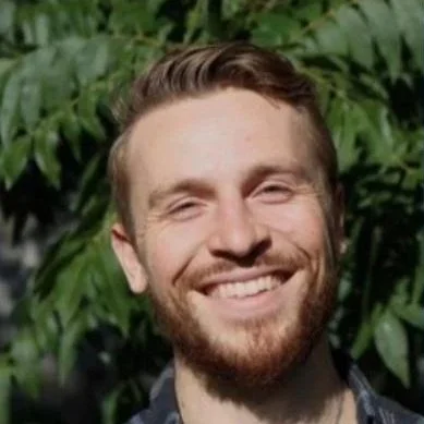 A man with short brown hair and a beard smiling outdoors with green foliage in the background.
