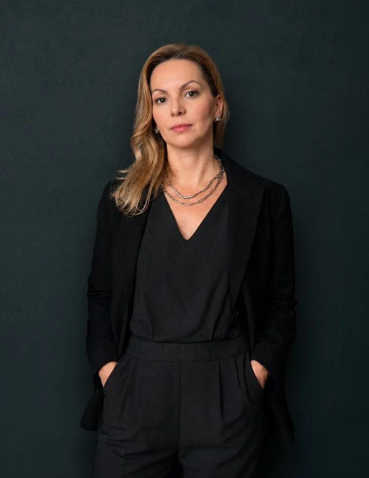A woman with light brown, wavy hair, wearing a black blazer and black pants, standing against a dark green background.
