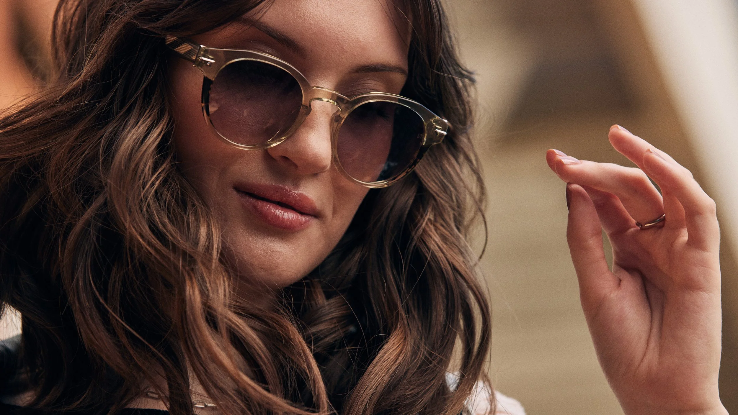 Close-up of a woman wearing sunglasses with a tortoiseshell frame, wavy brown hair, and a subtle smile, touching her sunglasses with her right hand.