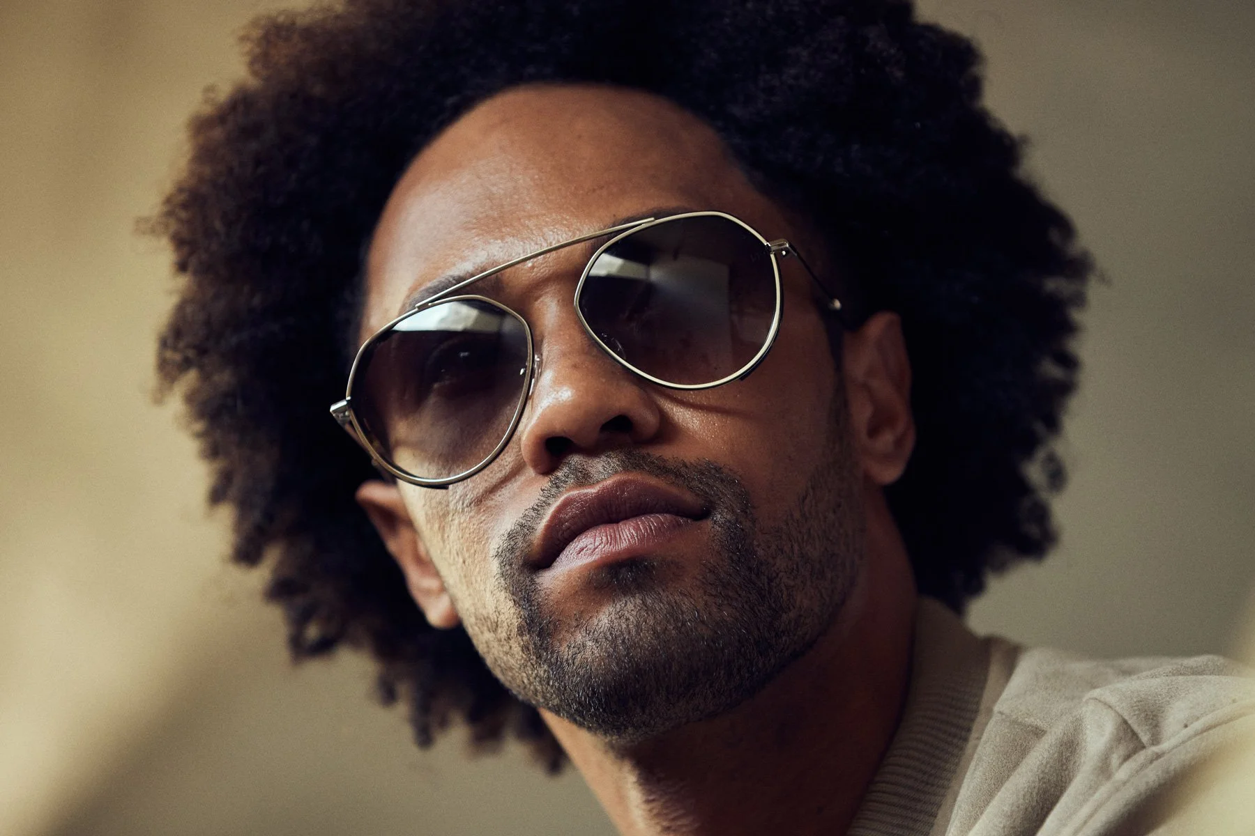 Close-up of a man with a dark complexion, wearing large aviator sunglasses with dark lenses and a light-colored shirt, with an Afro hairstyle, looking slightly upwards against a plain background.