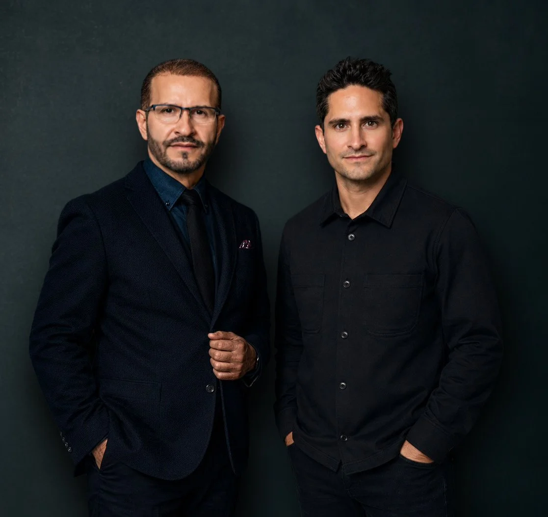 Two men dressed in black suits and shirts standing against a dark background.