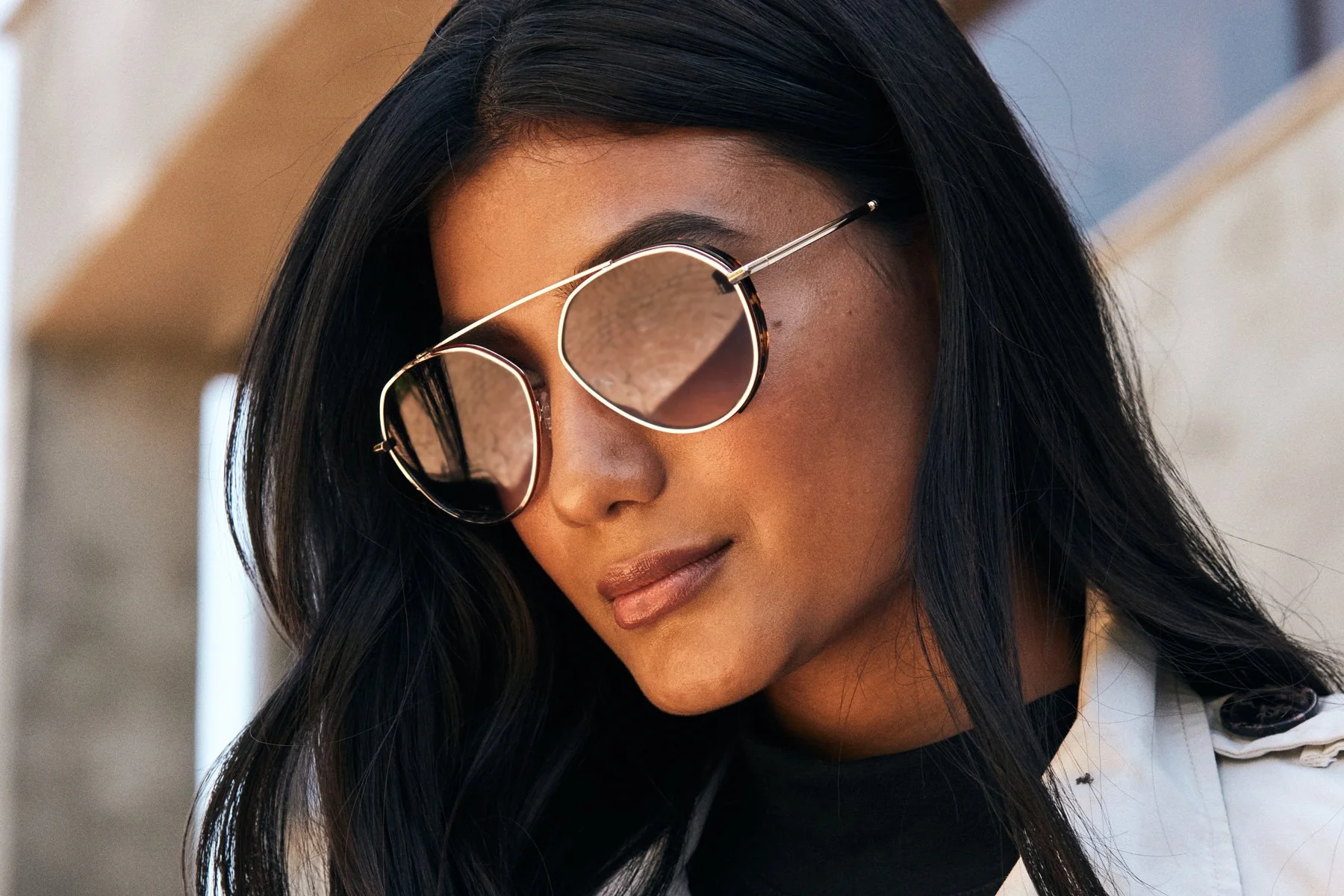 Close-up of a woman with long black hair, wearing stylish sunglasses with metal gold frames, and a white jacket.