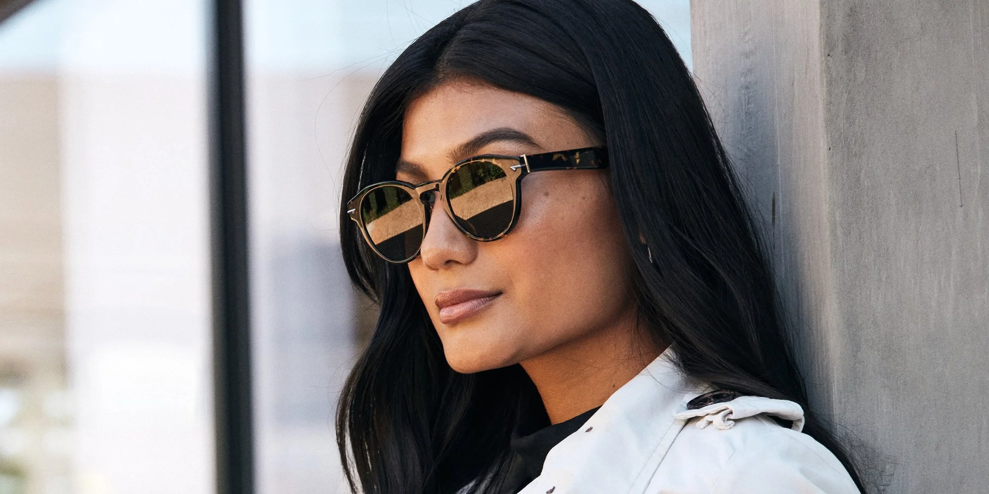 A woman with long black hair wearing stylish acetate sunglasses leaning against a wall outdoors.
