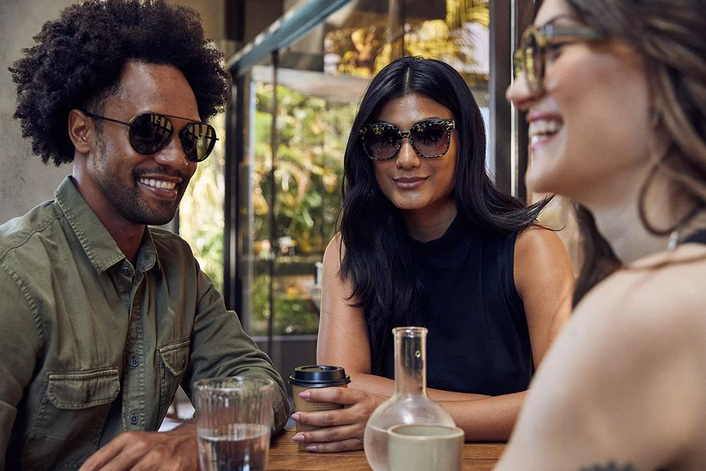 Three friends sitting at a table, smiling and talking outdoors, wearing sunglasses and casual clothes, with a glass of water and a bottle on the table.