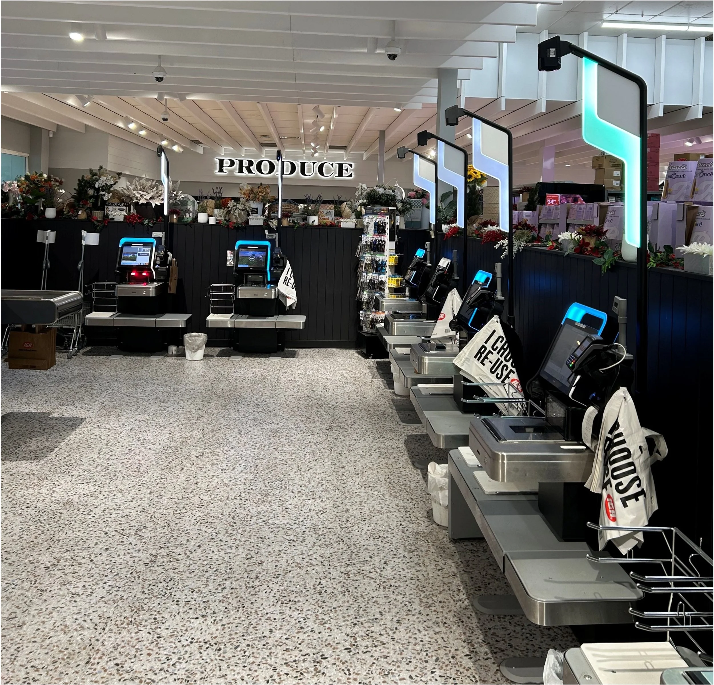 Grocery checkout area with self-service check-in kiosks, shopping bags, and floral decorations, located near an aisle with produce.