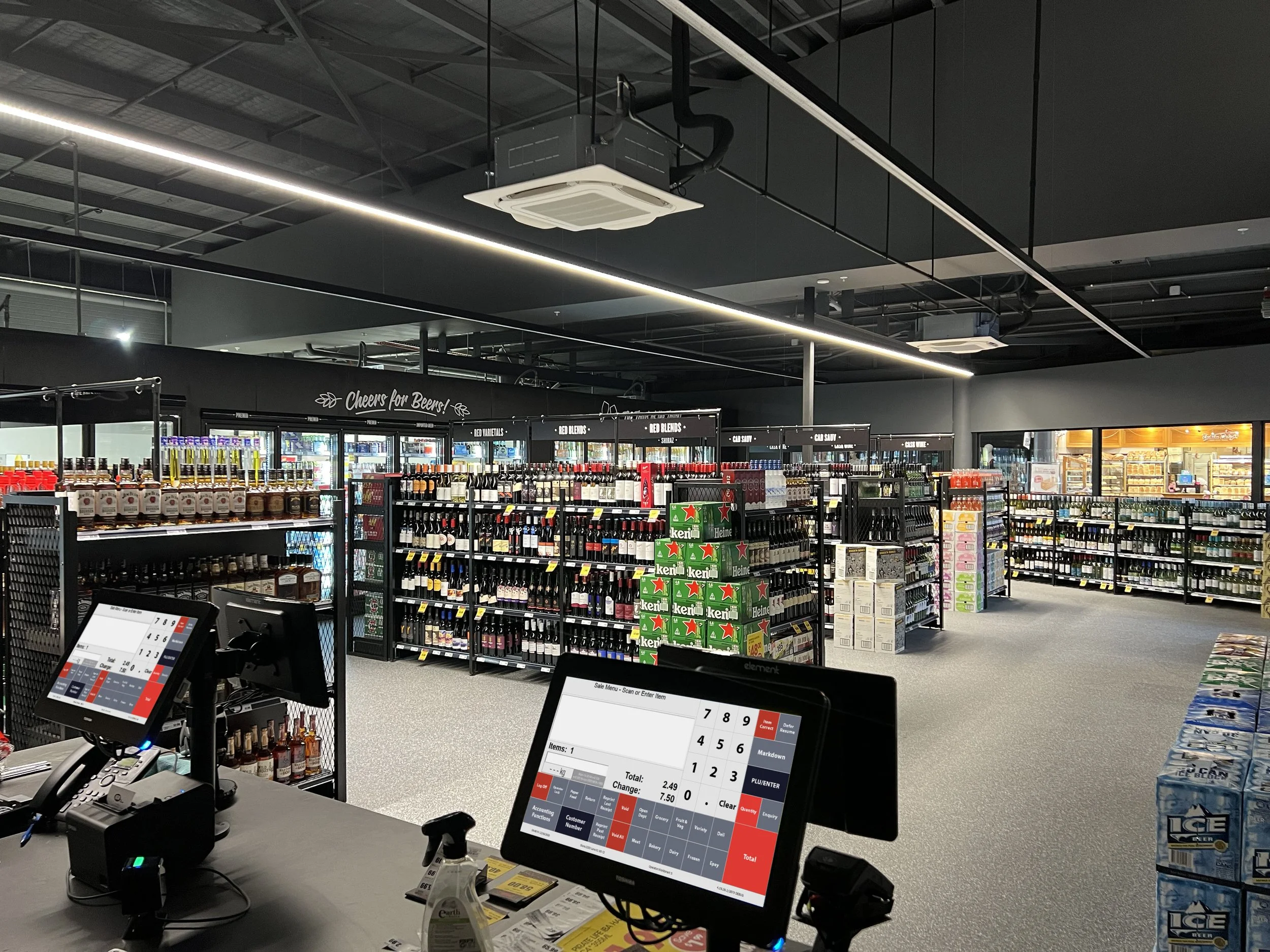 View of checkout counters with cash registers and point of sale screens at a liquor store, shelves stocked with wine and liquor bottles, and a sign reading 'Cheers for Beers!'