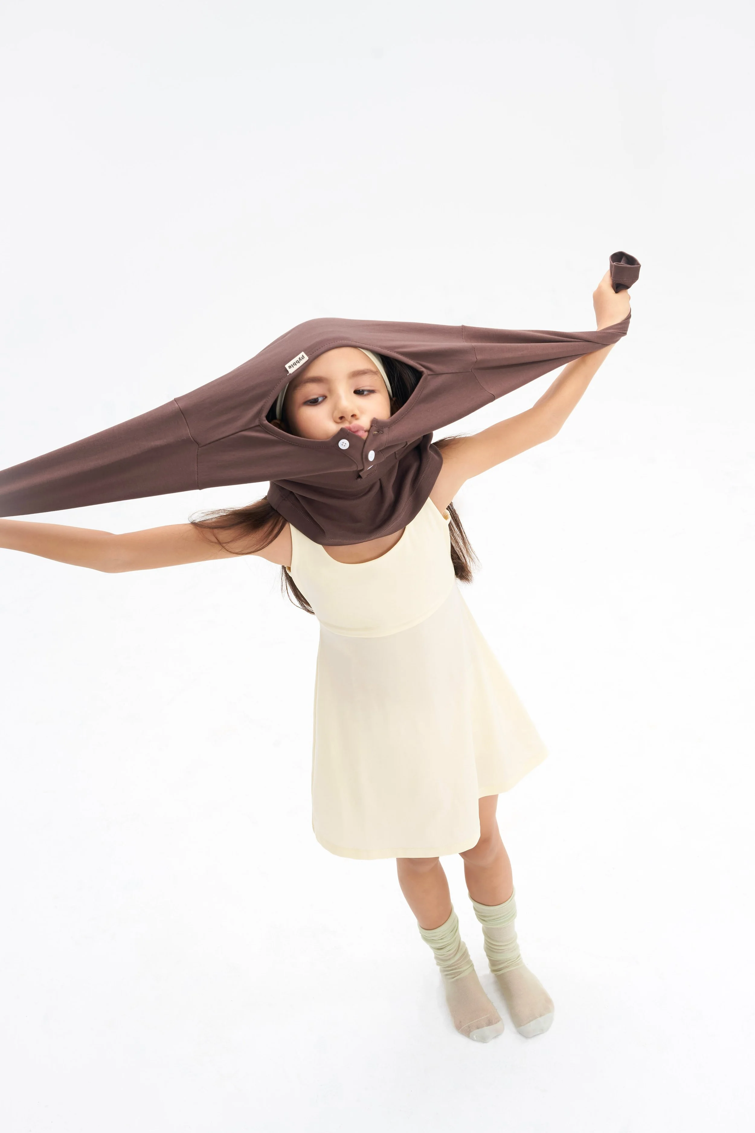 Young girl in a white dress and beige socks, wearing a brown hoodie with a hood that extends into a cape, standing against a white background.