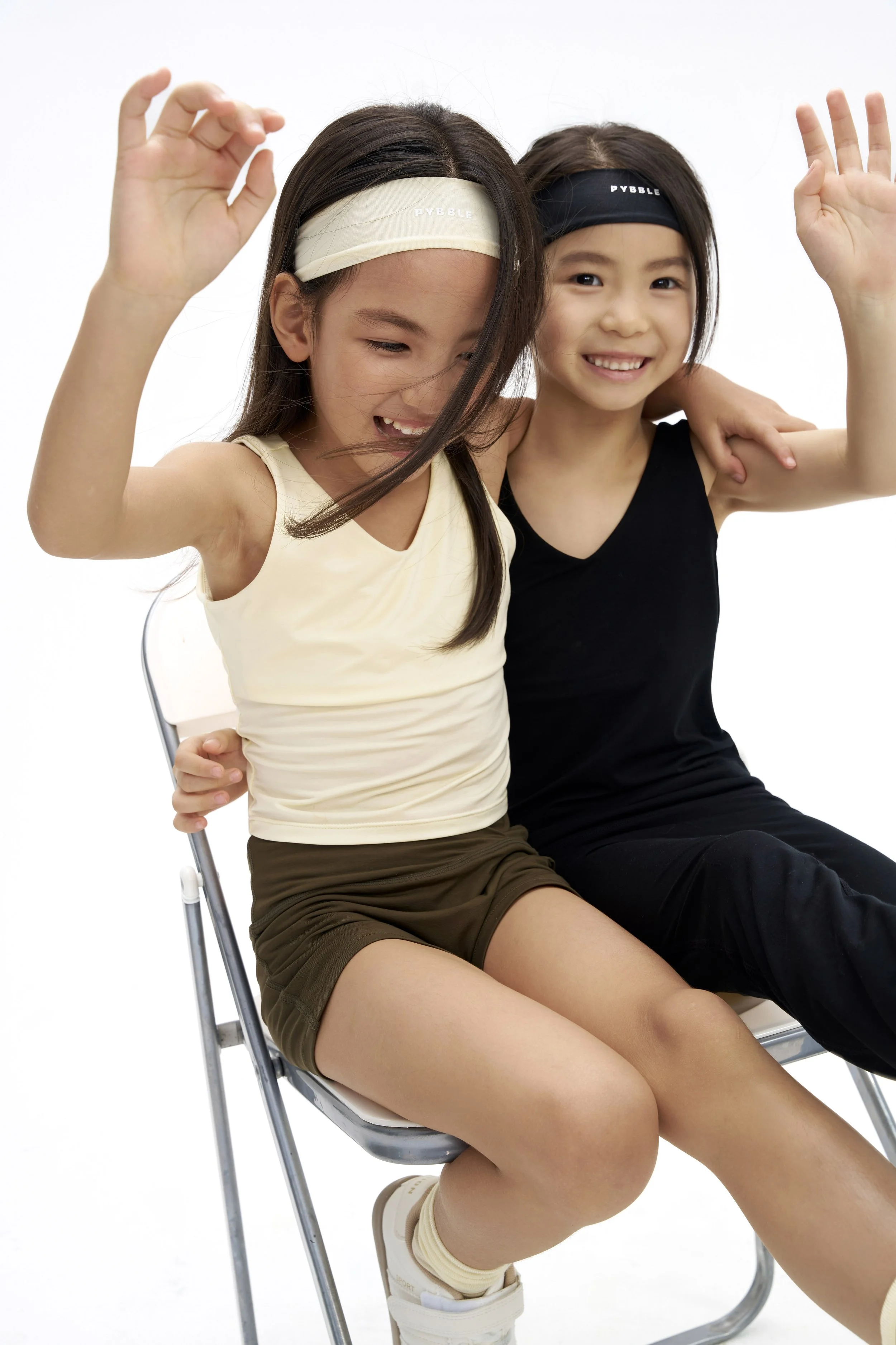 Two young girls sitting on a metal chair, smiling and waving, wearing sports headbands and athletic clothing, against a plain white background.