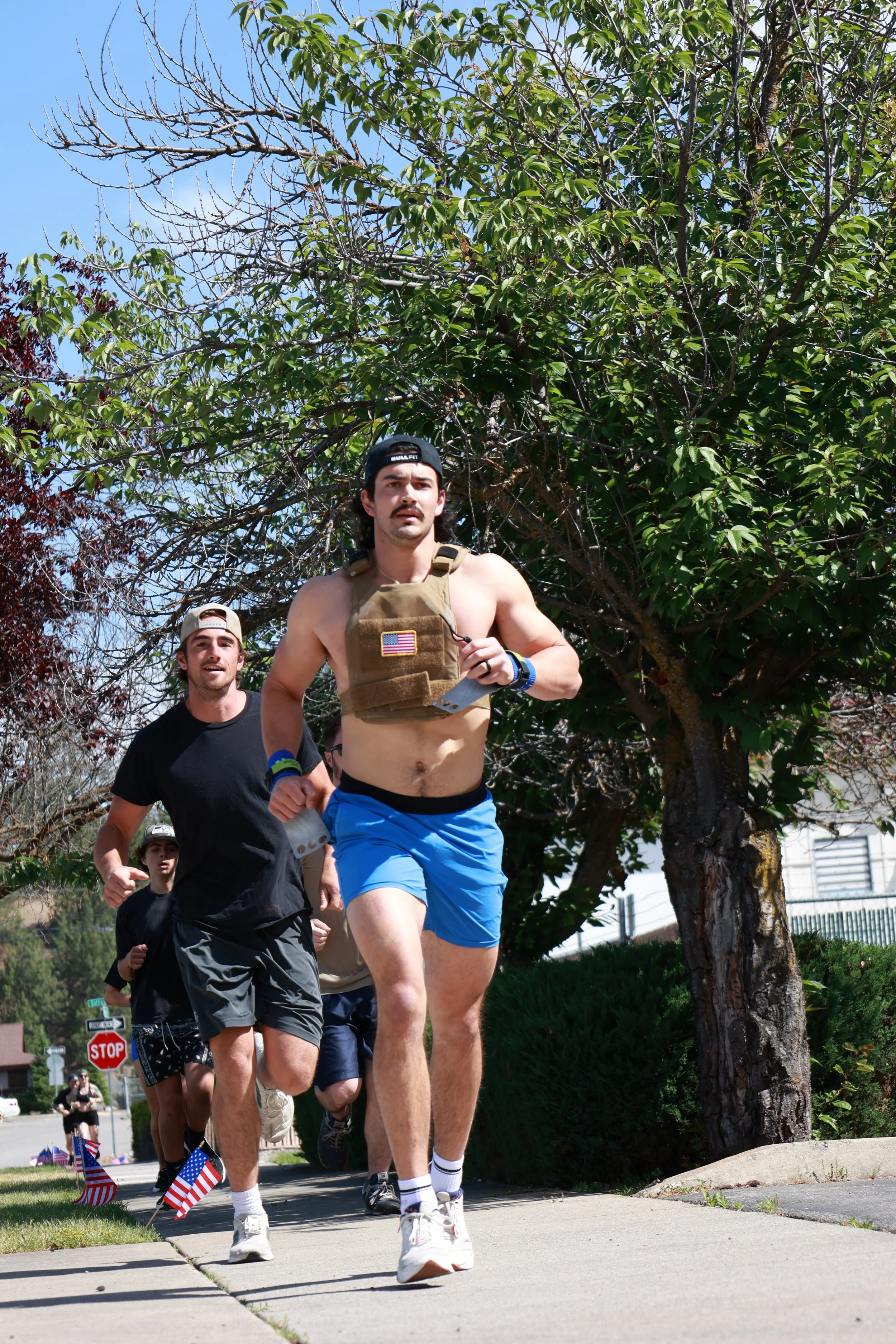 A group of people running outdoors on a sidewalk, with small American flags planted in the grass along the path, some holding flags, during daytime with a clear sky, trees, and traffic signs visible in the background.