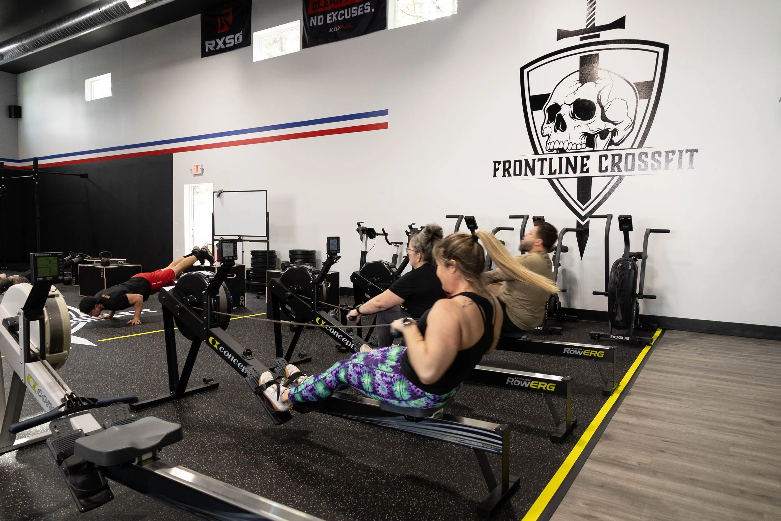 People exercising on rowing machines inside a gym with a large skull and shield logo and the words 'Frontline CrossFit' on the wall.