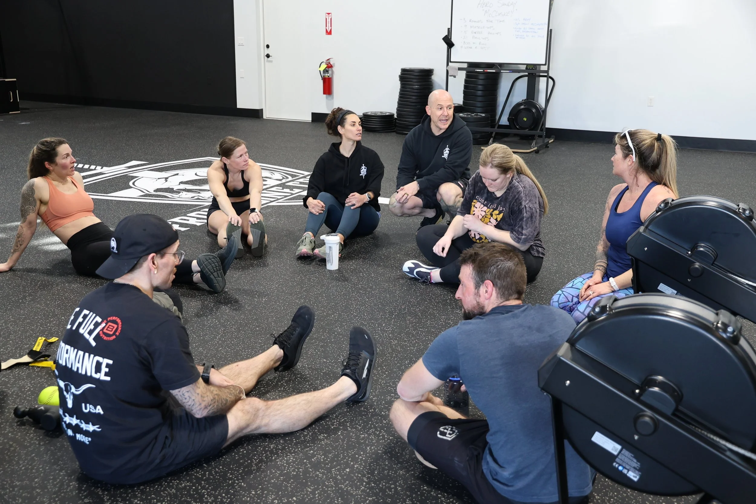 Group of people sitting on the gym floor in a circle, engaging in a post-workout stretching or discussion, with gym equipment and a whiteboard in the background.