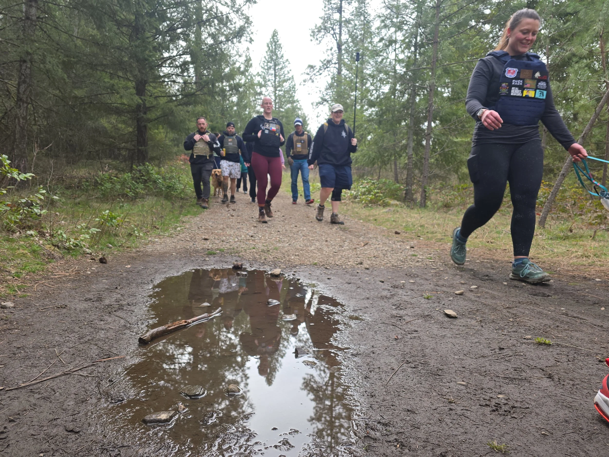 A group of people trail running in a forest, following a dirt path with small puddles and surrounded by tall green trees.