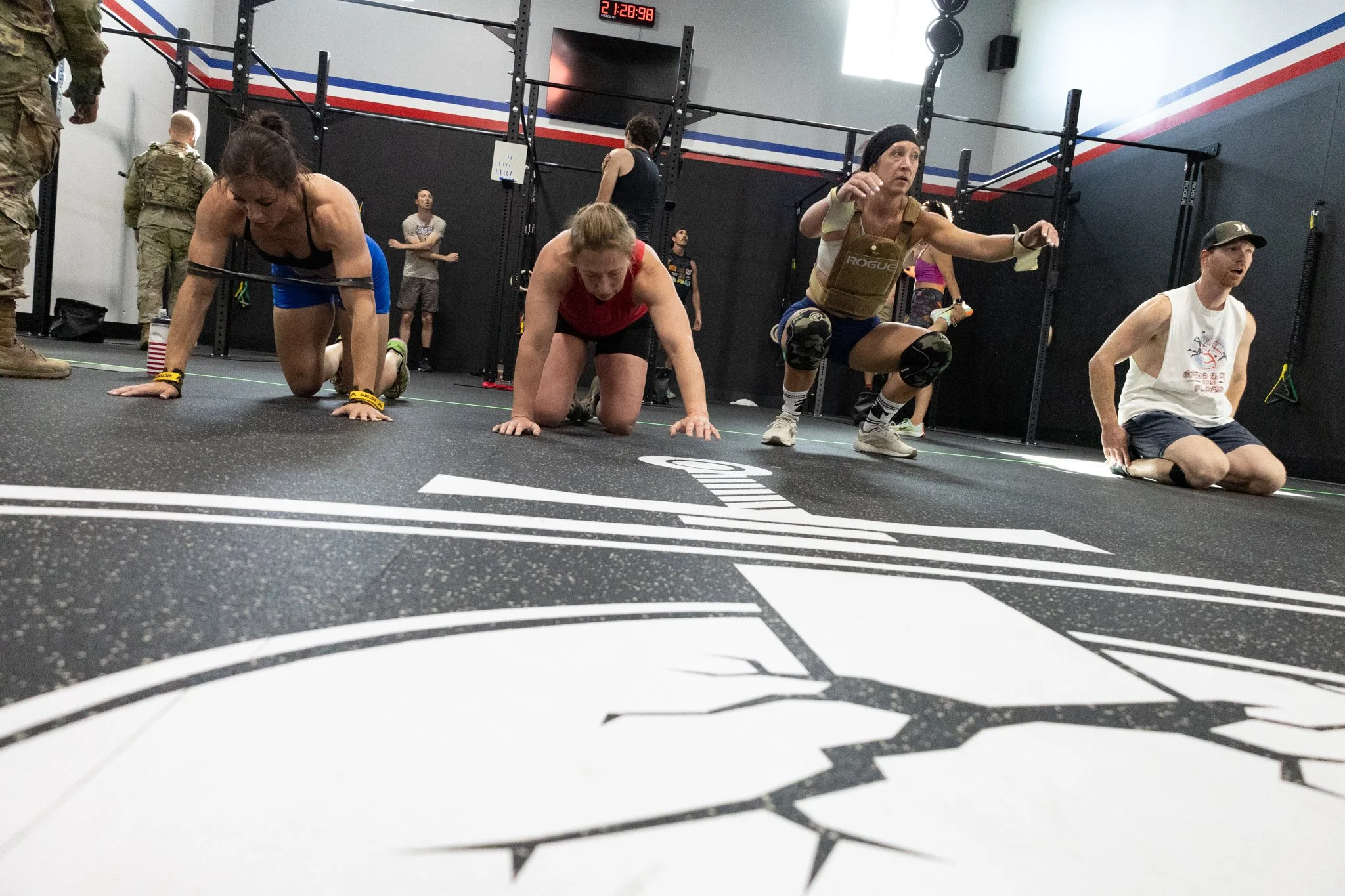 Women performing push-ups on a black gym floor with a white and black logo, in a CrossFit gym with black walls, training racks, and a digital timer display.