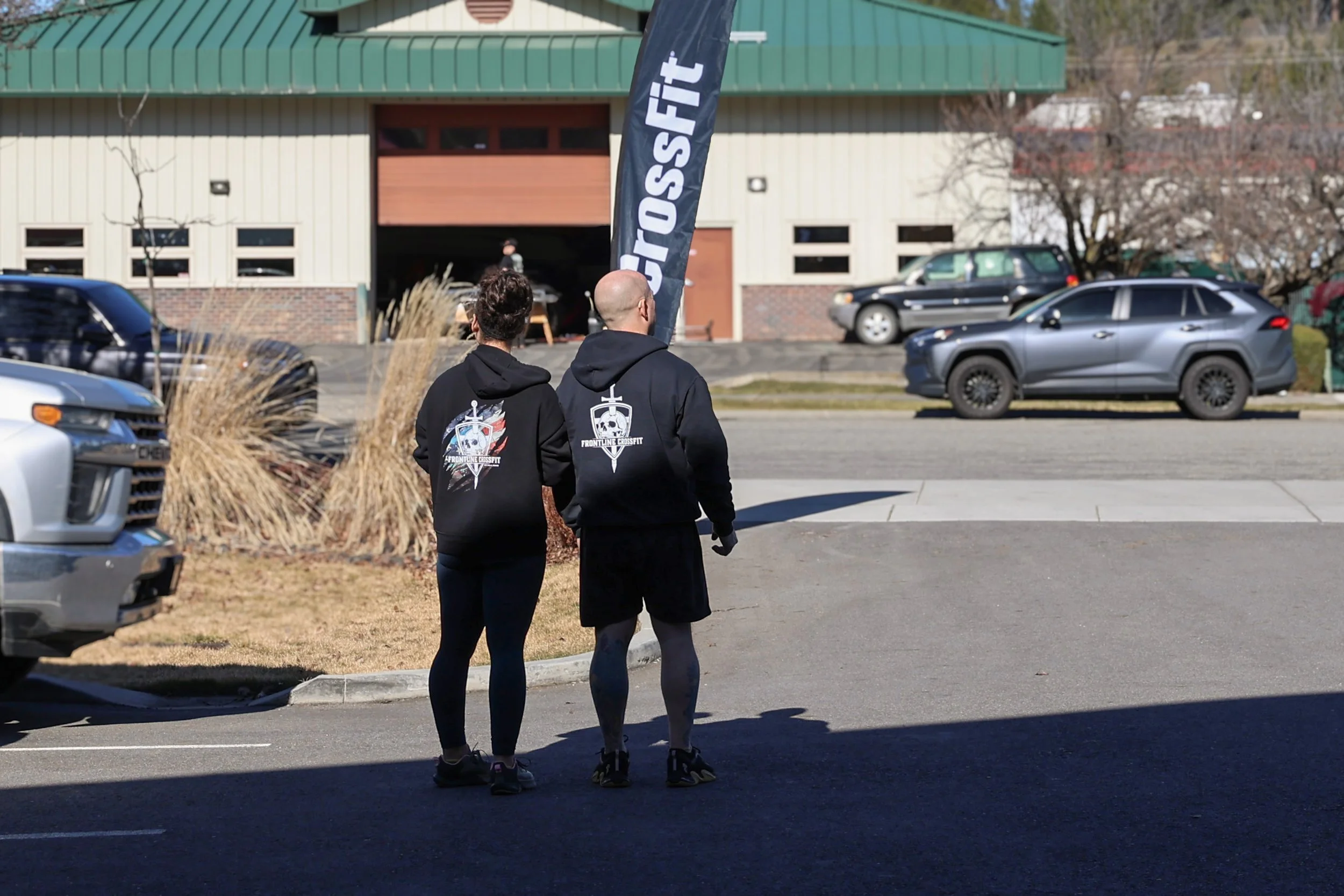 Couple standing in a parking lot just outside Frontline CrossFit, wearing black 'Frontline CrossFit' hoodies and waiting for members to find their way to the gym.