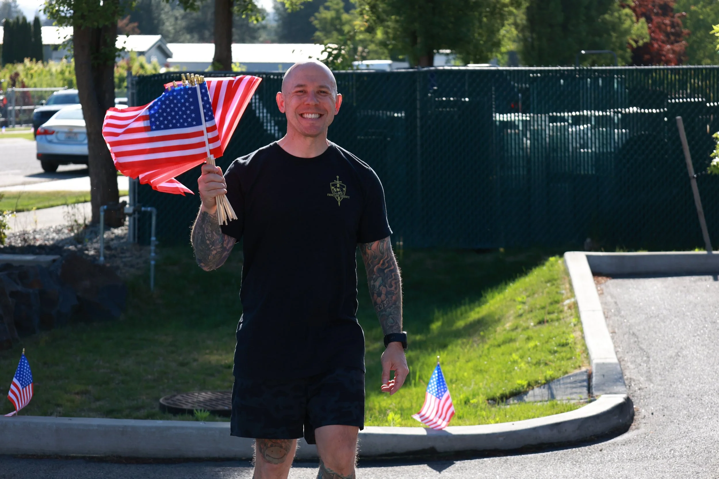 A patriotic man with tattoos on his arm holding multiple American flags outdoors on a sunny day honoring those who made the ultimate sacrifice on Memorial Day.