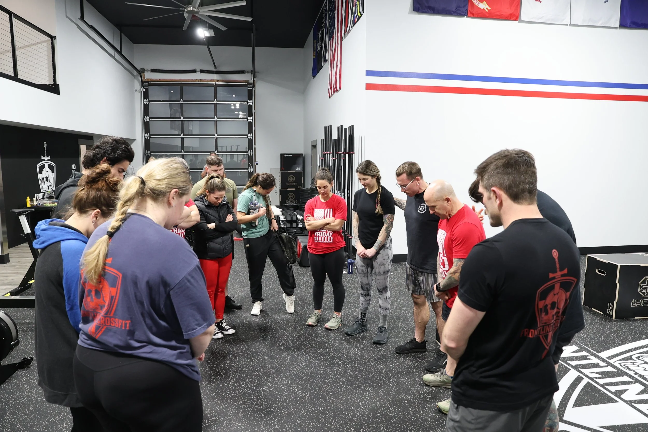 A group of people gathered in a gym, standing in a circle with their heads bowed and eyes closed. They appear to be participating in a moment of reflection or prayer.