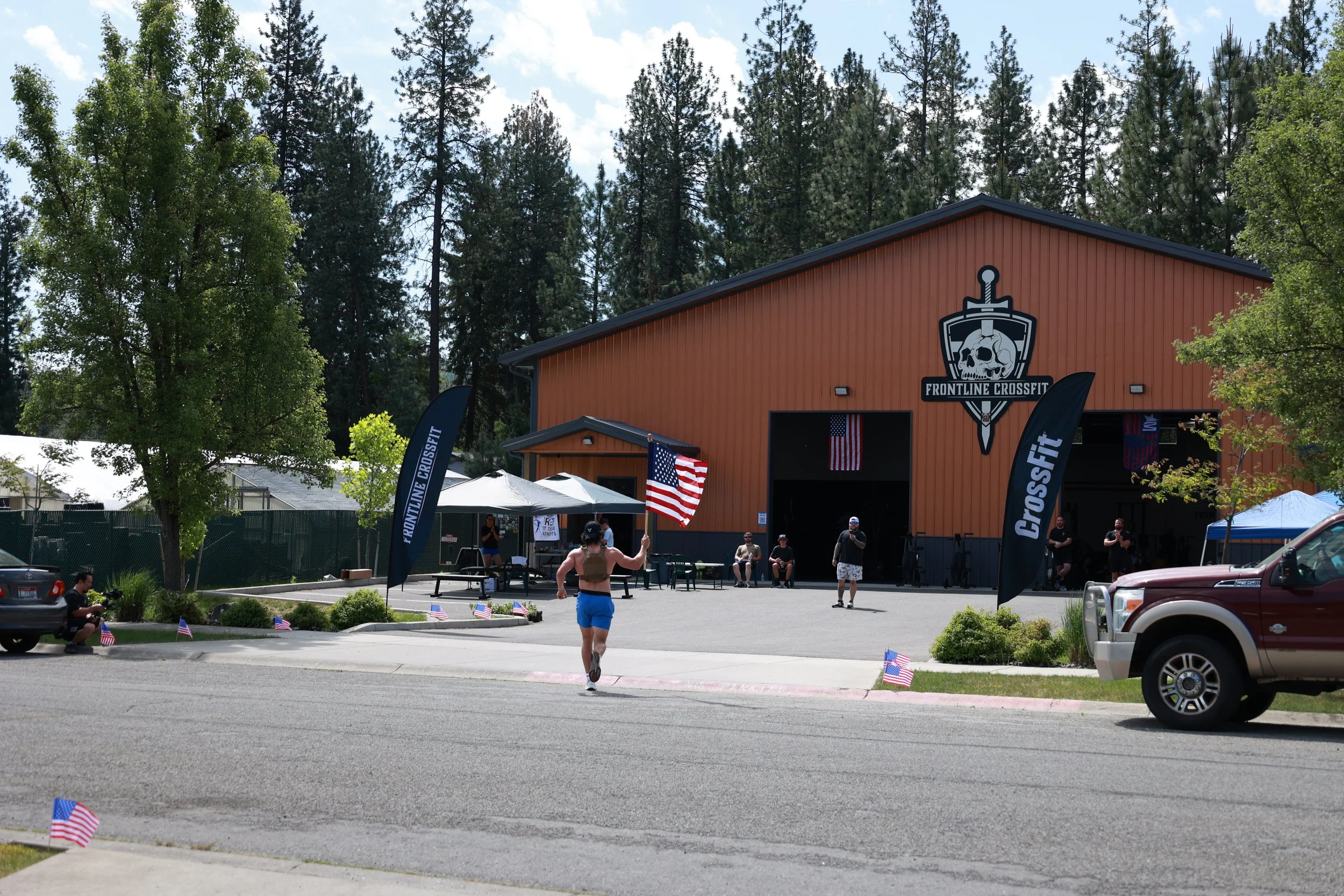 An outdoor scene at Frontline CrossFit gym with people participating in a workout, American flags lined along the parking lot, and the gym building with a large CrossFit logo featuring a skull and sword on the front.