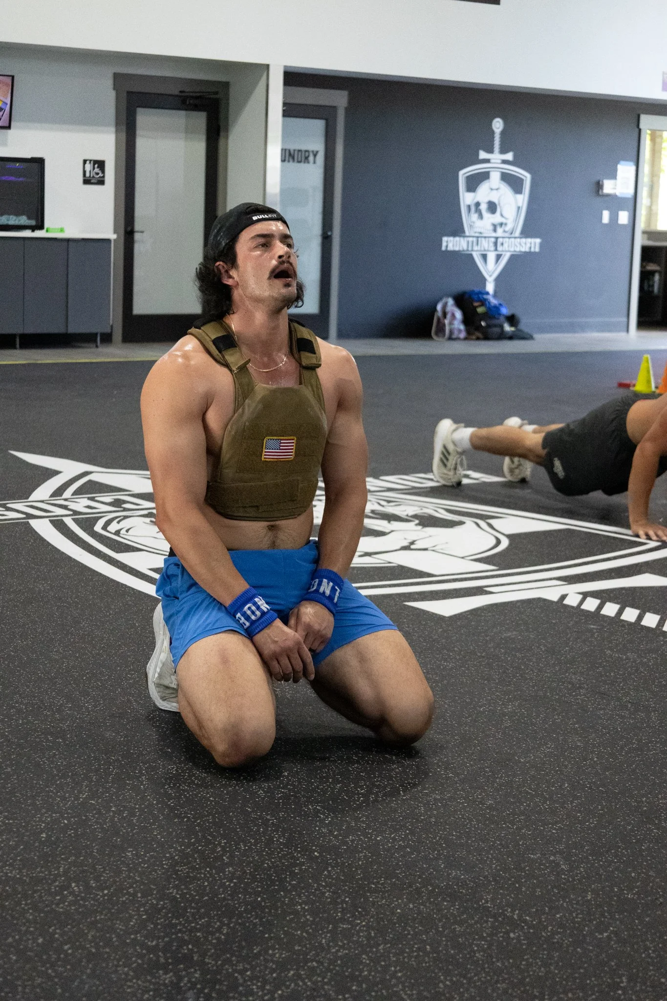 A man kneeling on a gym floor in front of a CrossFit gym logo, wearing a military-style vest, blue shorts, a black cap, and wrist wraps, with another person exercising in the background.