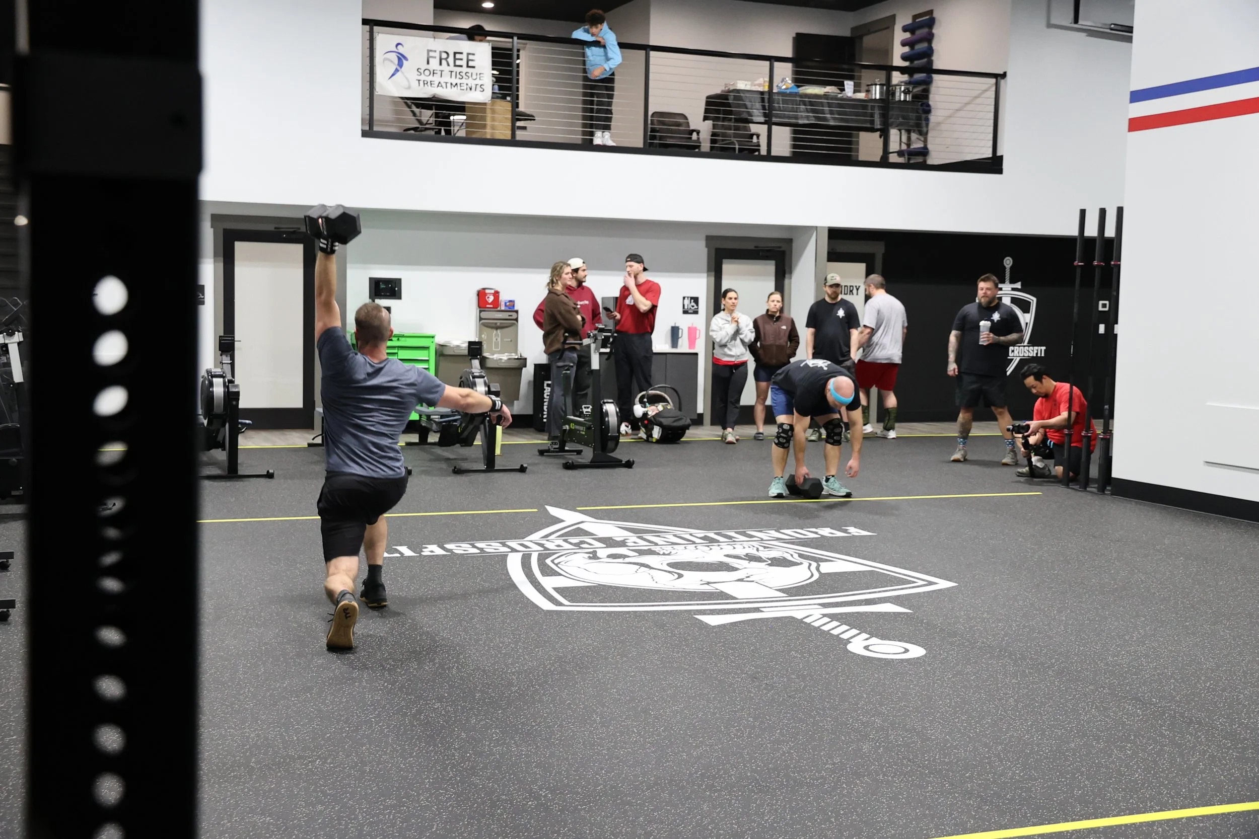 People working out inside the Frontline CrossFit gym, one person lifting weights, others talking and observing, second floor with someone leaning over railing, signs for free soft tissue treatments and a medical professional.
