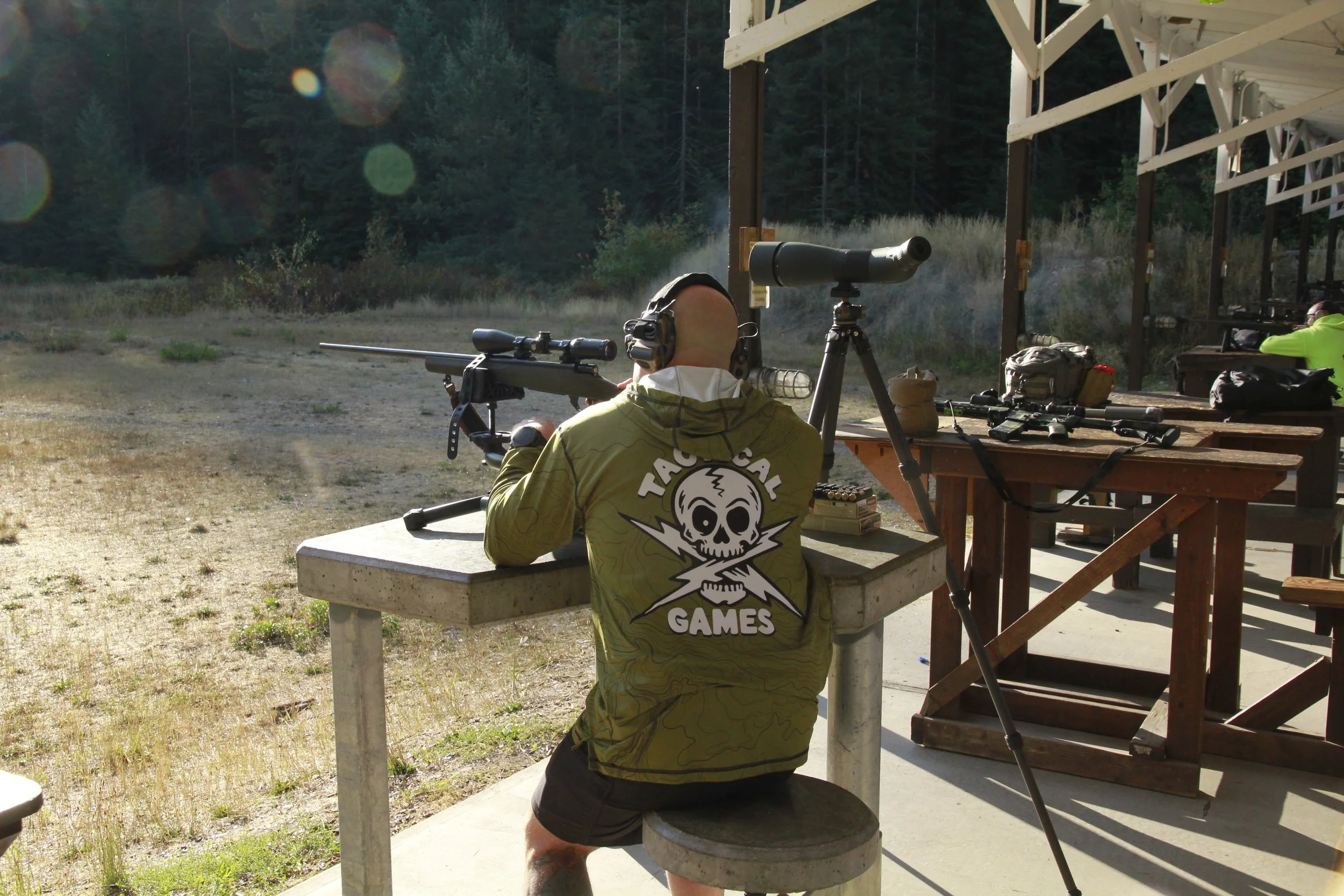 A person with a shaved head wearing a yellow jacket and headphones, sitting at a shooting range with a rifle and spotting scope, surrounded by trees and outdoors.