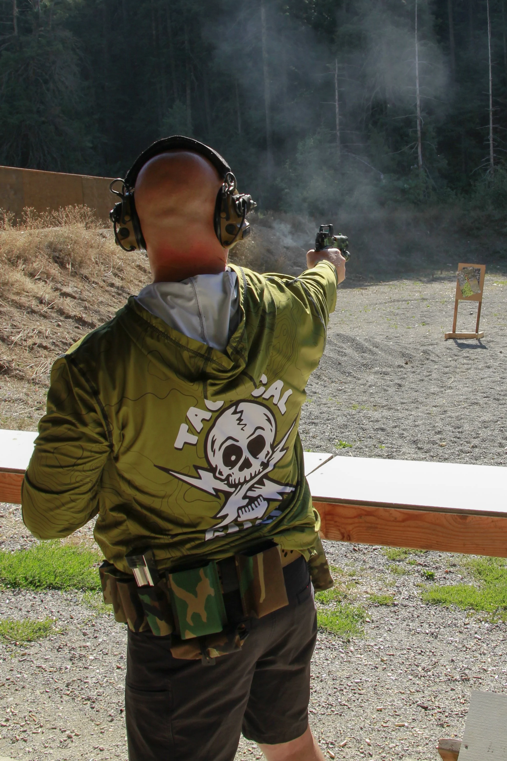 A person shooting a handgun at an outdoor shooting range. The person is wearing hearing protection and a camouflage jacket with a skull logo. There is a target set up in the background surrounded by trees.