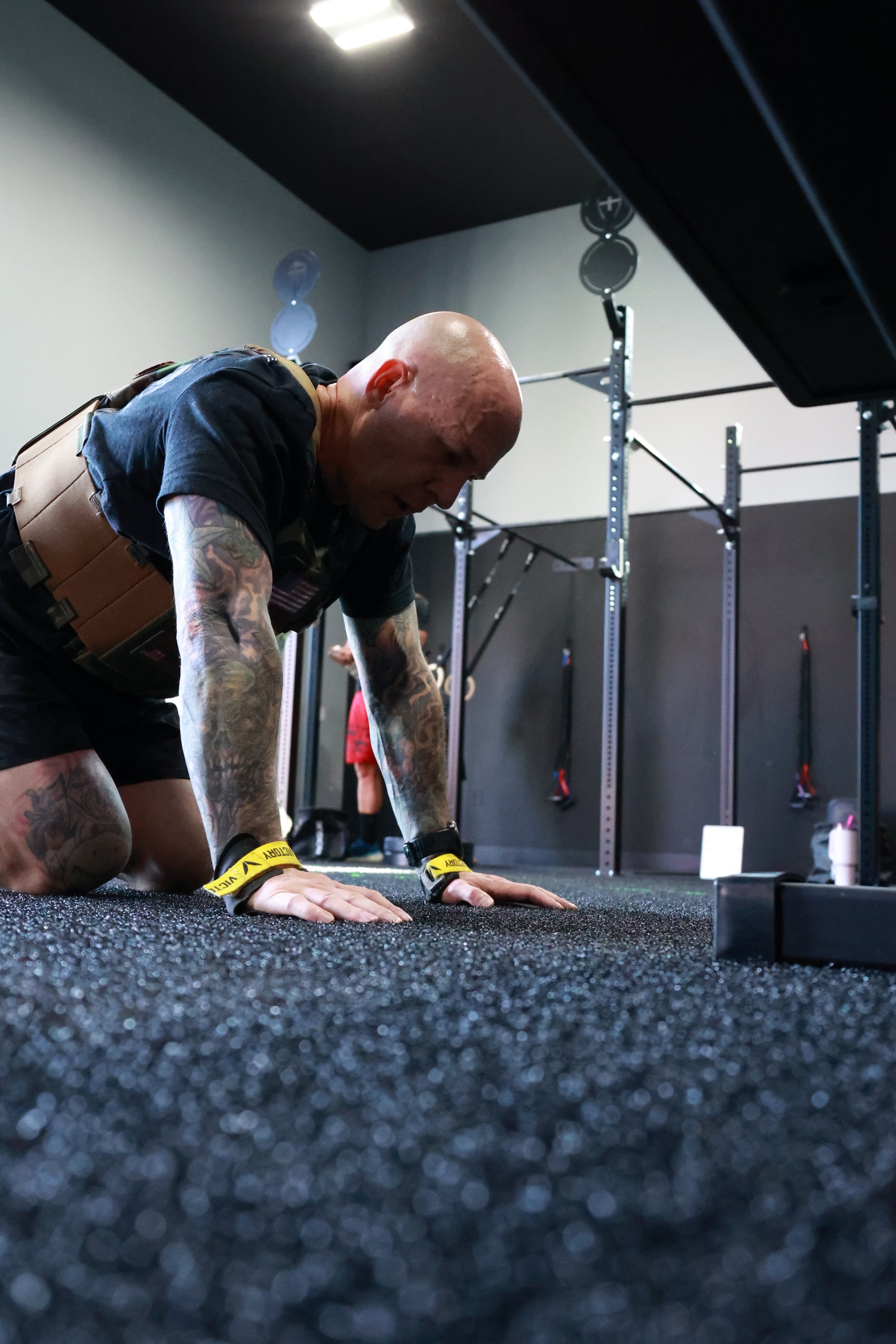 A man with tattoos on his arms and a bald head is doing a push-up on a black gym mat in a fitness studio with black walls and gym equipment in the background.