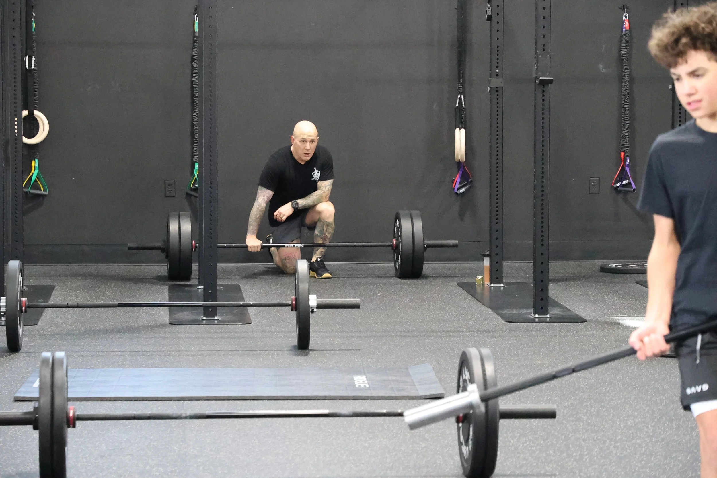 A man with tattoos and a shaved head is preparing to lift a barbell at a gym, with various gym equipment and wall-mounted exercise bands in the background. A person with curly hair is partially visible on the right side of the image.