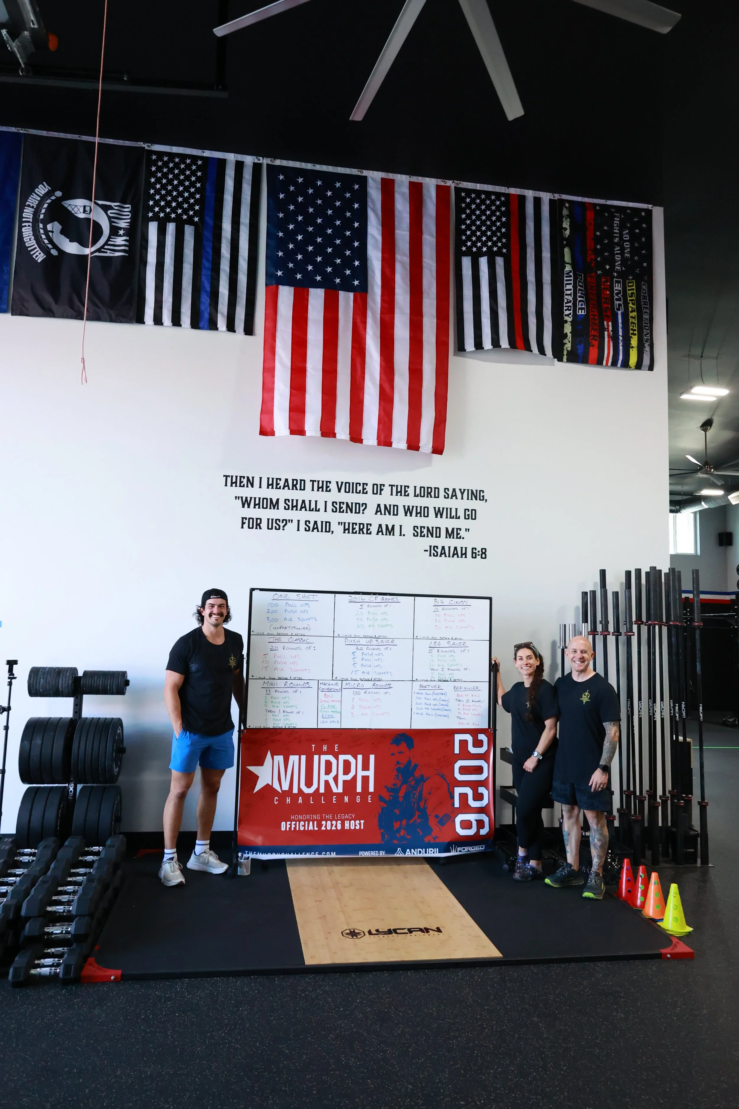 Group of four people standing in a gym with fitness flags hanging on the wall, including American flags and others with police, military, and fitness slogans, and a large red banner for the Murphy Challenge 2020. One person is on the left side, and three others are on the right, all smiling. There are weightlifting and exercise equipment around.
