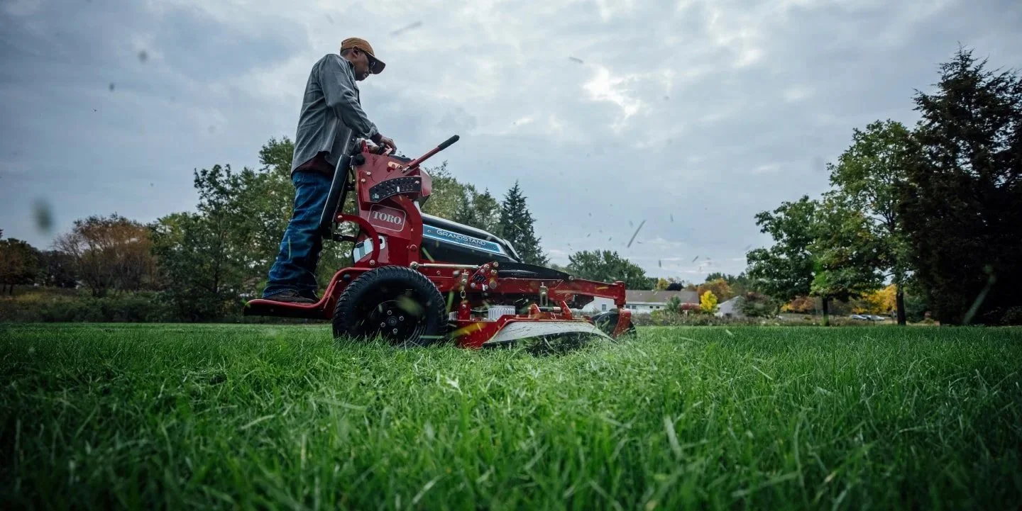 A person mowing a lawn with a red riding mower on a grassy field with trees and cloudy sky in the background.
