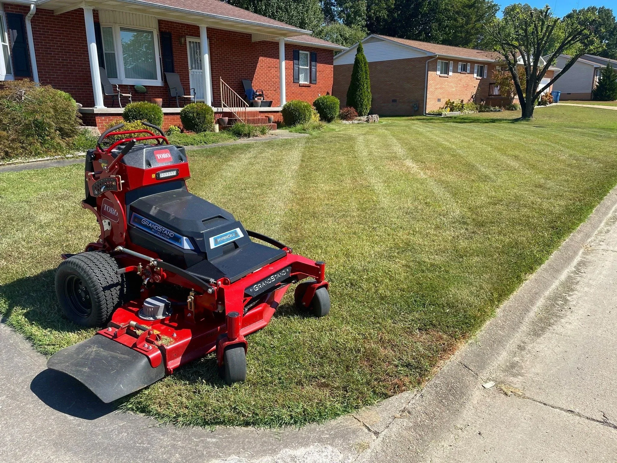 A red riding lawn mower on the edge of a grass lawn in front of a brick house with a porch and chairs. There are bushes and trees in the yard, and other houses are visible in the background.