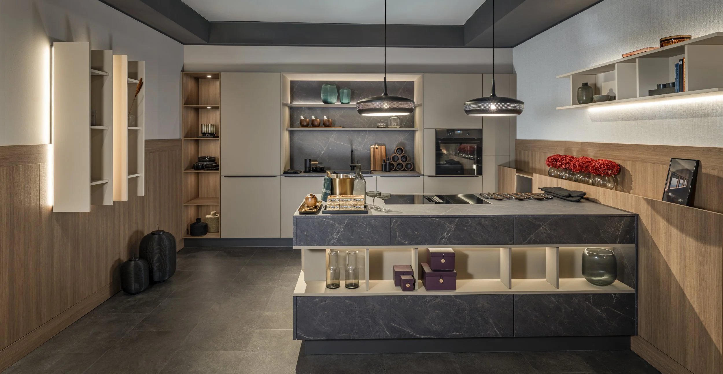 Cuisine moderne avec armoires grises, étagères en bois, plan de travail en marbre noir, décorations en verre et céramique, suspensions modernes, et décoration florale rouge.