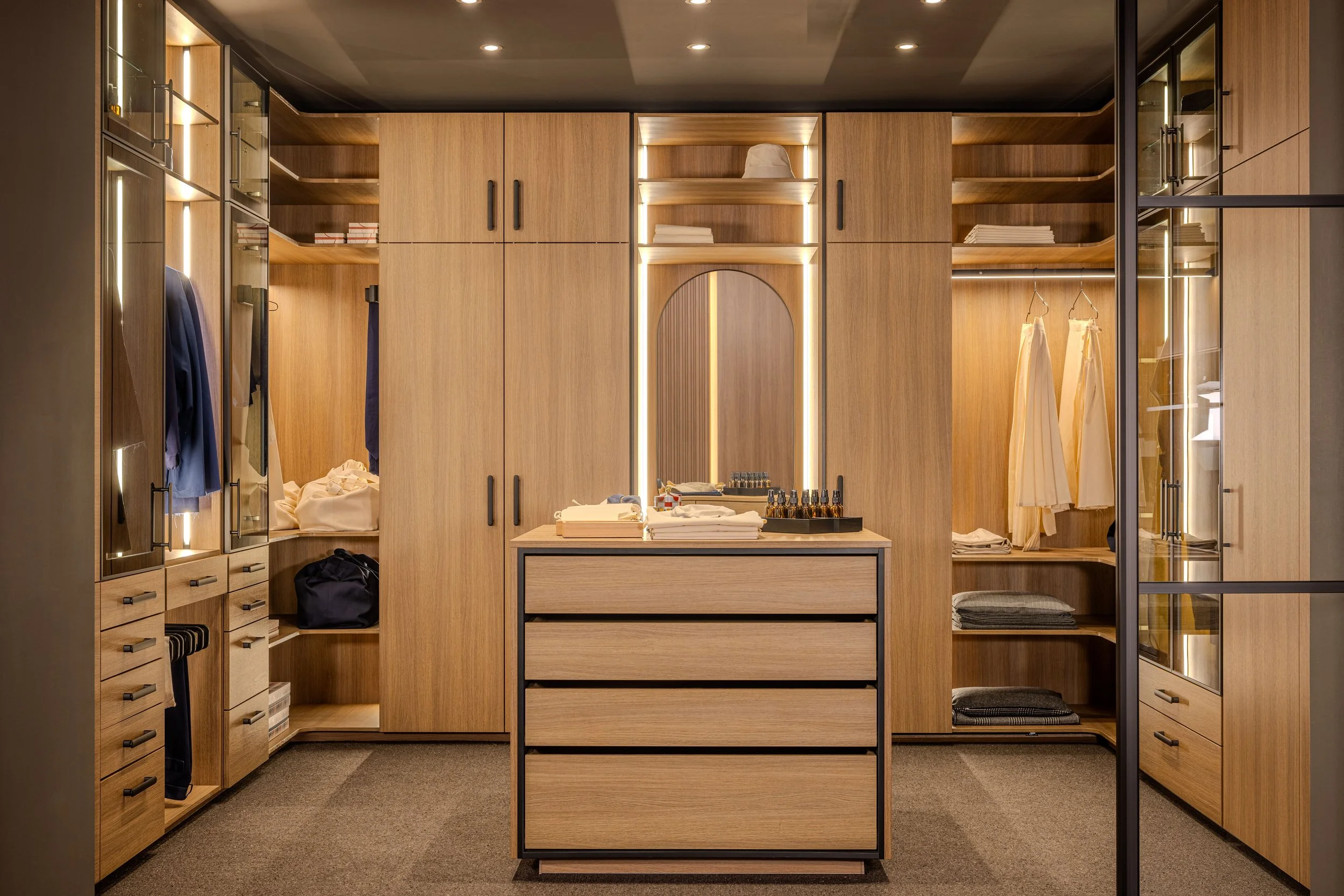 Une garde-robe en bois avec beaucoup d'étagères et de compartiments, contenant des vêtements, des livres et des accessoires, avec un miroir rond encadré de lumières LED et une table en bois en premier plan.