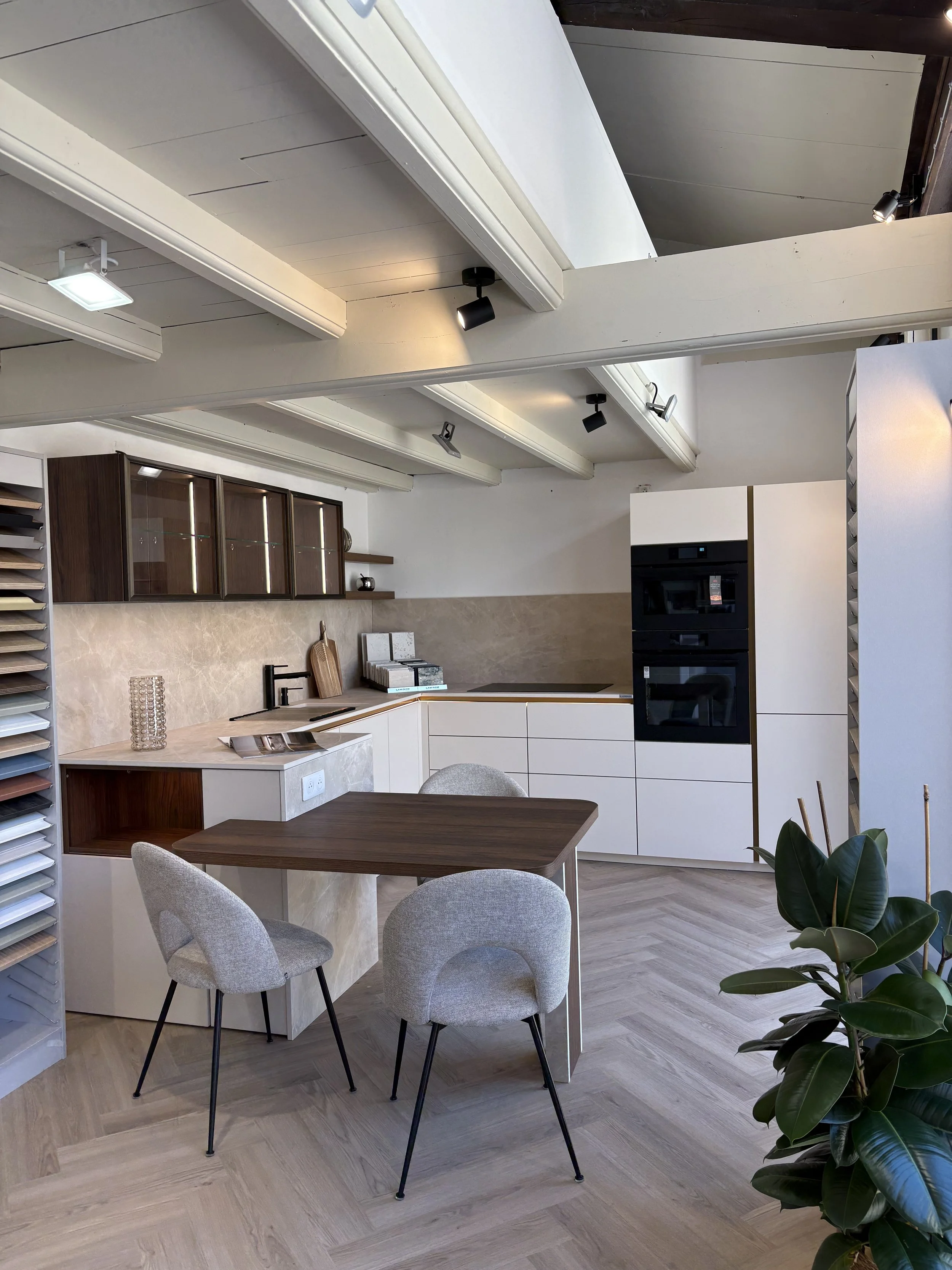 Une cuisine moderne avec une table en bois et quatre chaises en tissu gris clair, un plan de travail avec des meubles en bois foncé et blanc, et un mur en beige avec des appareils électroménagers intégrés. Il y a aussi une plante verte dans le coin droit