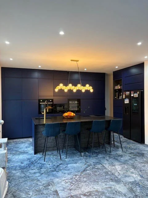 Cuisine moderne avec un îlot central, chaises bleues, éclairage suspendu doré, murs bleus foncés, réfrigérateur, et décorations sur les étagères.