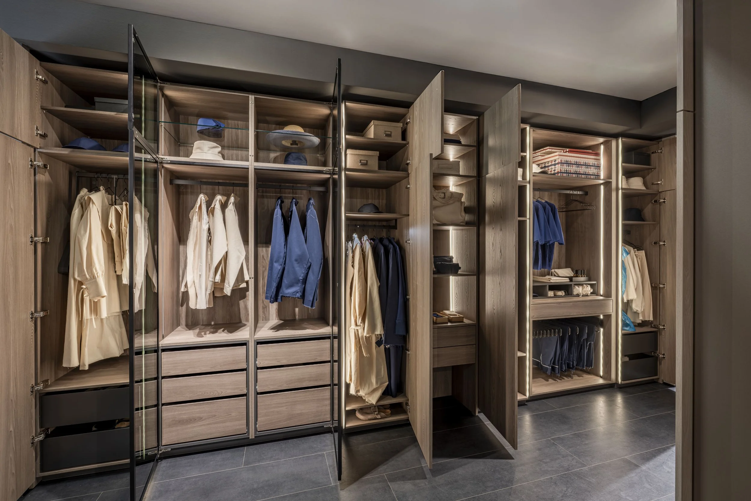Une armoire en bois avec des portes ouvertes contenant des vêtements, des casquettes, des boîtes et des chapeaux, dans une pièce moderne avec un sol en carreaux noirs.