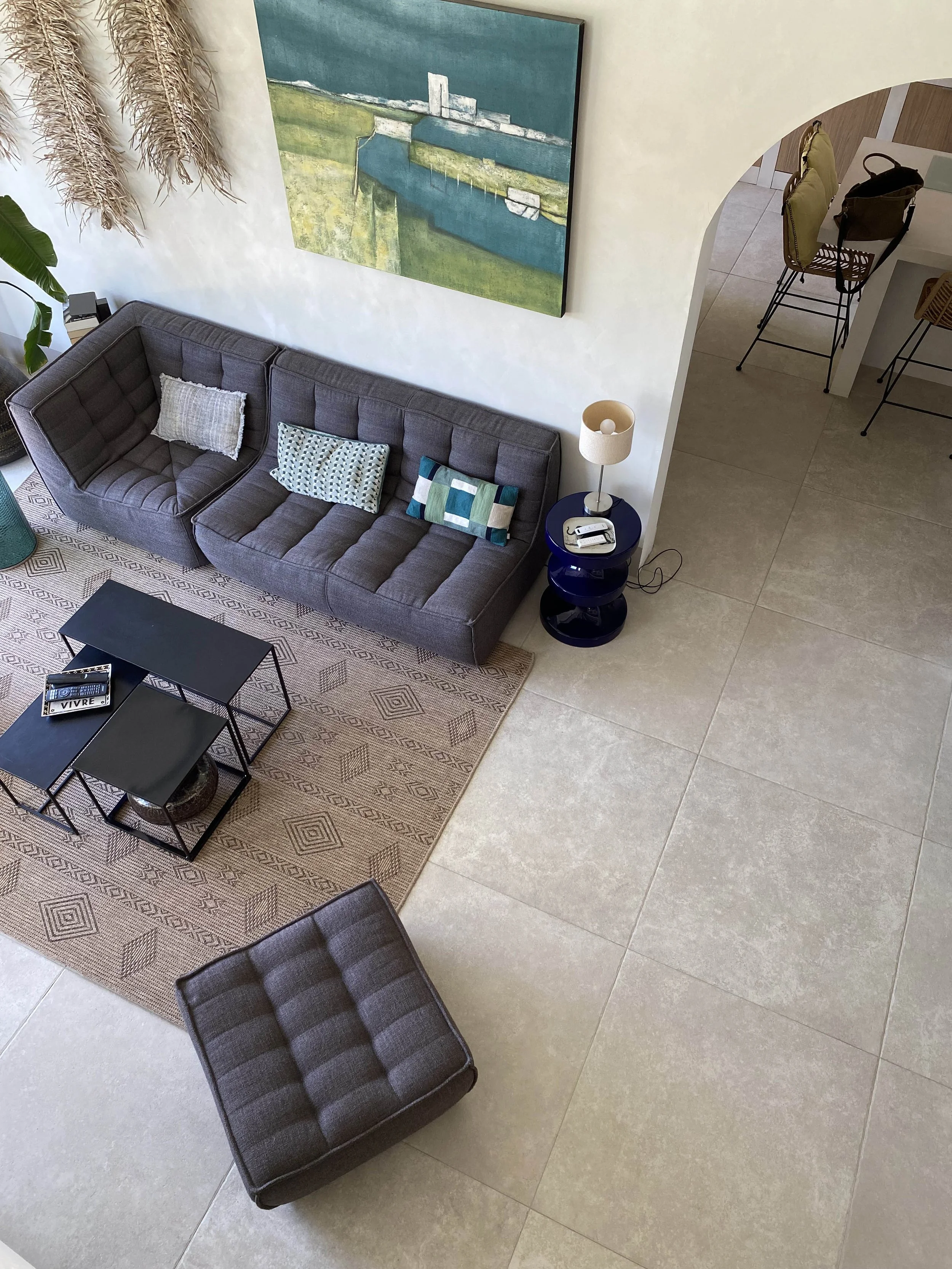 Salon avec un canapé gris, une table basse noire, un tapis beige à motifs, peinture moderne sur le mur et un lampadaire à côté du canapé. Il y a aussi quelques chaises avec manteaux sur le comptoir visible à droite.