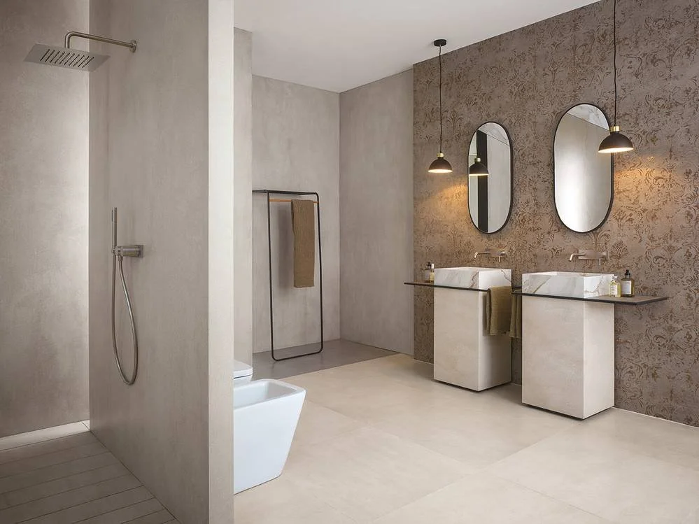 Une salle de bain moderne avec deux lavabos, deux miroirs ovales, un lavabo en marbre, des lampes suspendues, un mur avec un motif délicat, une douche avec une tête de douche au plafond, une étagère à serviettes, un porte-serviettes, et un sol en car