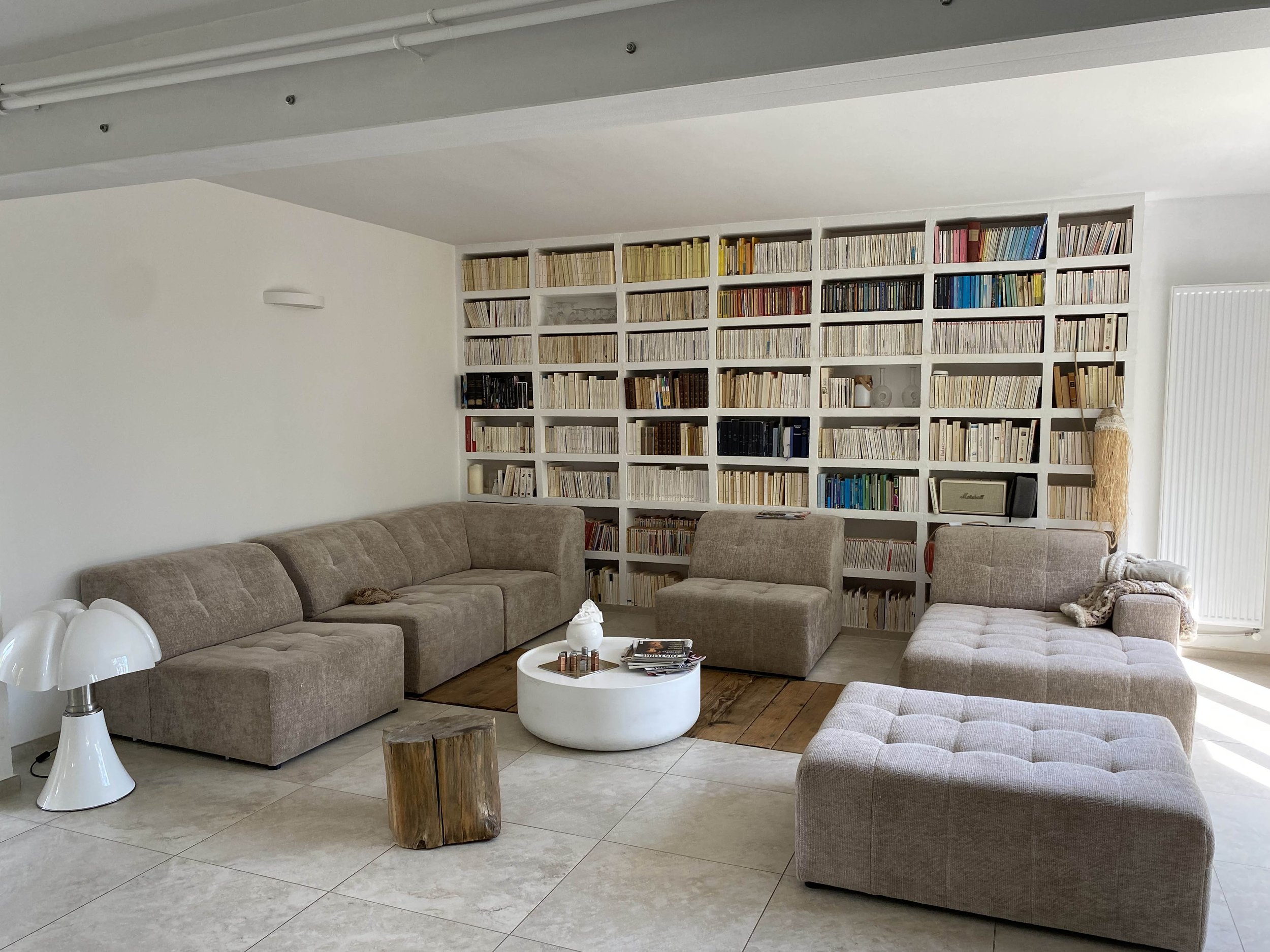 Un salon avec un grand mur de bibliothèque rempli de livres, un canapé en forme de L, une table basse blanche ronde, une lampe blanche moderne, et des fauteuils beige clair dans une pièce lumineuse.