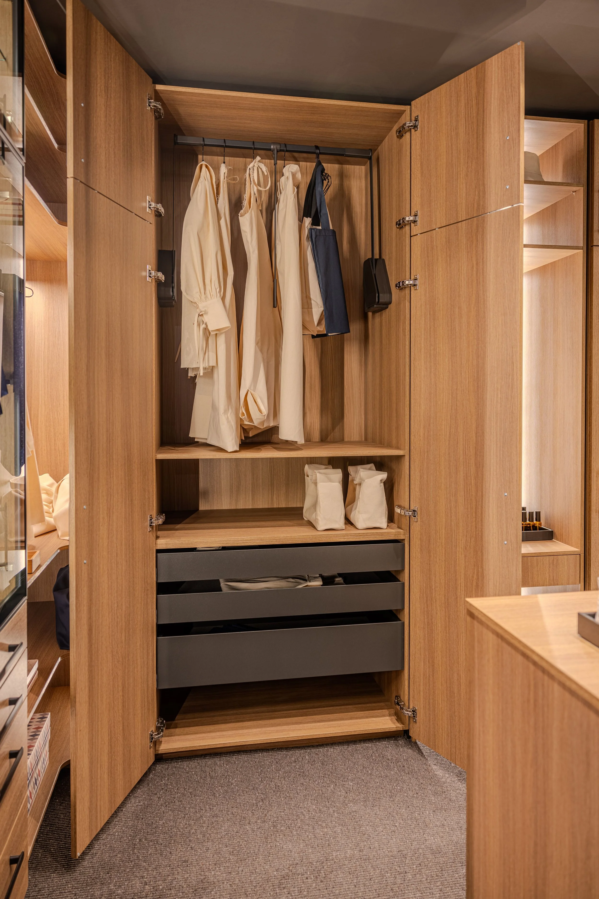 Un placard en bois avec des vêtements à l'intérieur, notamment des robes beige, et des sacs. Le sol est recouvert de moquette gris foncé.