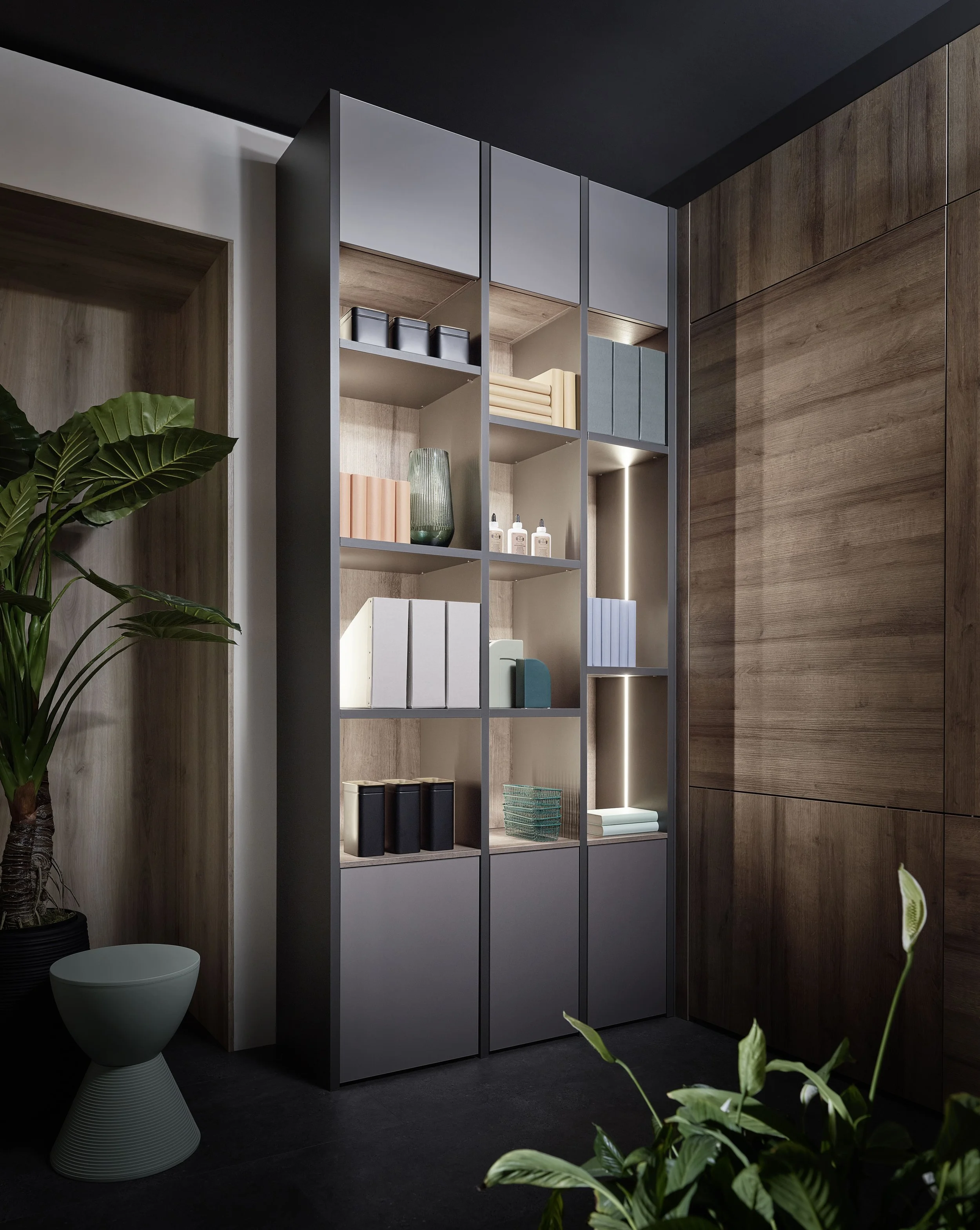 Une étagère moderne en bois clair avec des compartiments de rangement, située dans un intérieur avec des murs en bois foncé et en blanc, décorée avec des livres, des boîtes, un vase vert transparent et des bouteilles blanches.
