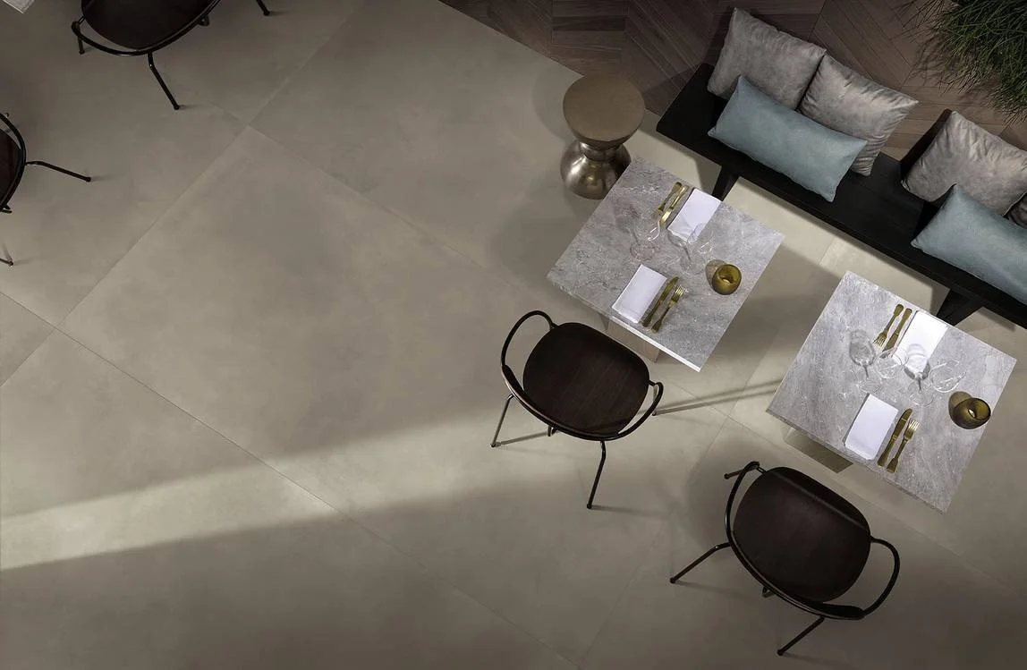 Vue d'une salle à manger moderne avec deux tables en marbre, quatre assiettes, couverts en or, verres et petits bols en or. Il y a un canapé gris avec des coussins bleus et gris, un petit meuble et des chaises noires en arrière-plan.