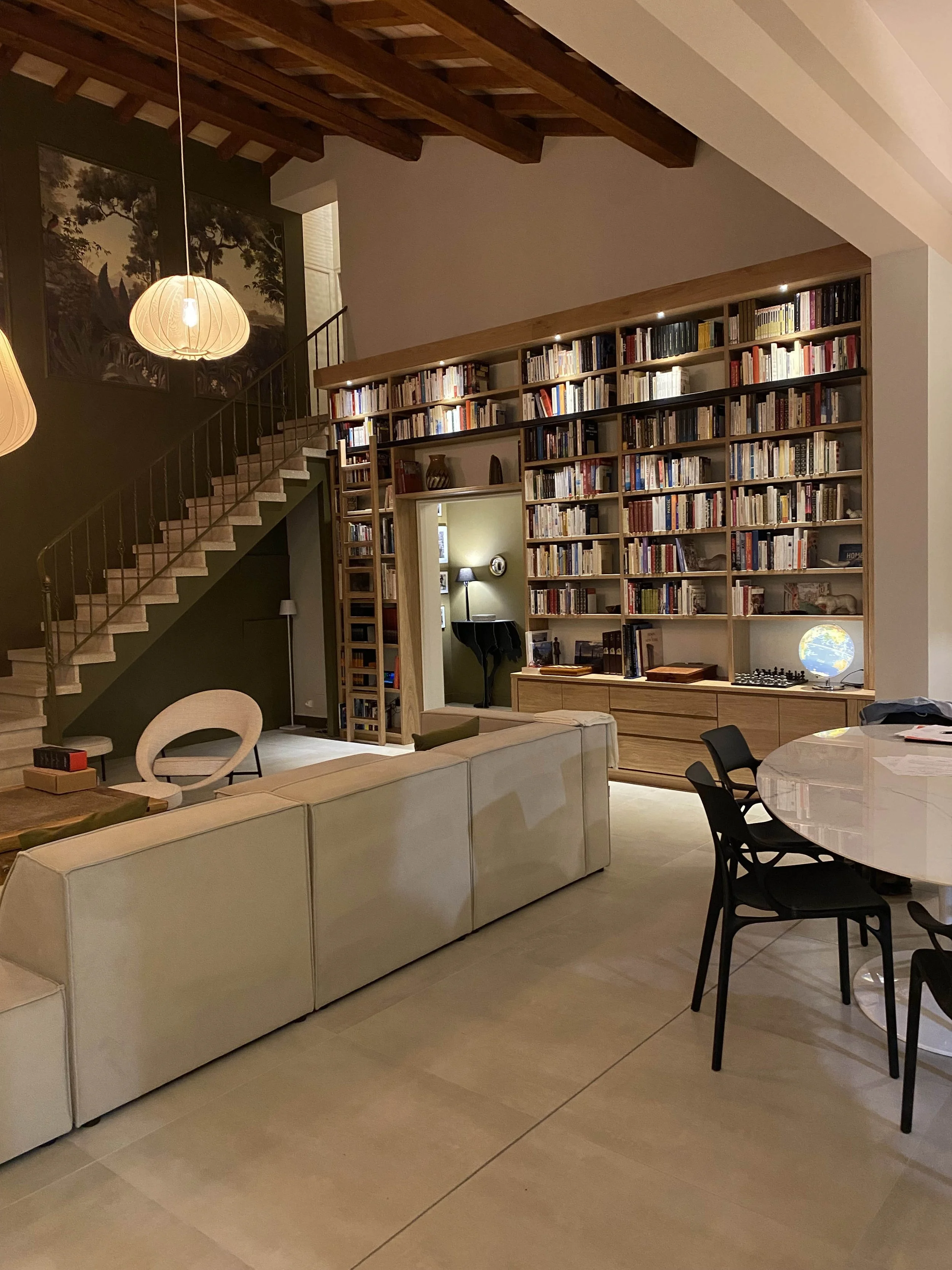 Salon avec canapé blanc, table ronde avec chaises noires, bibliothèque remplie de livres, globe terrestre lumineux, lampes suspendues, escalier en colimaçon, tableau de paysages dans le mur.